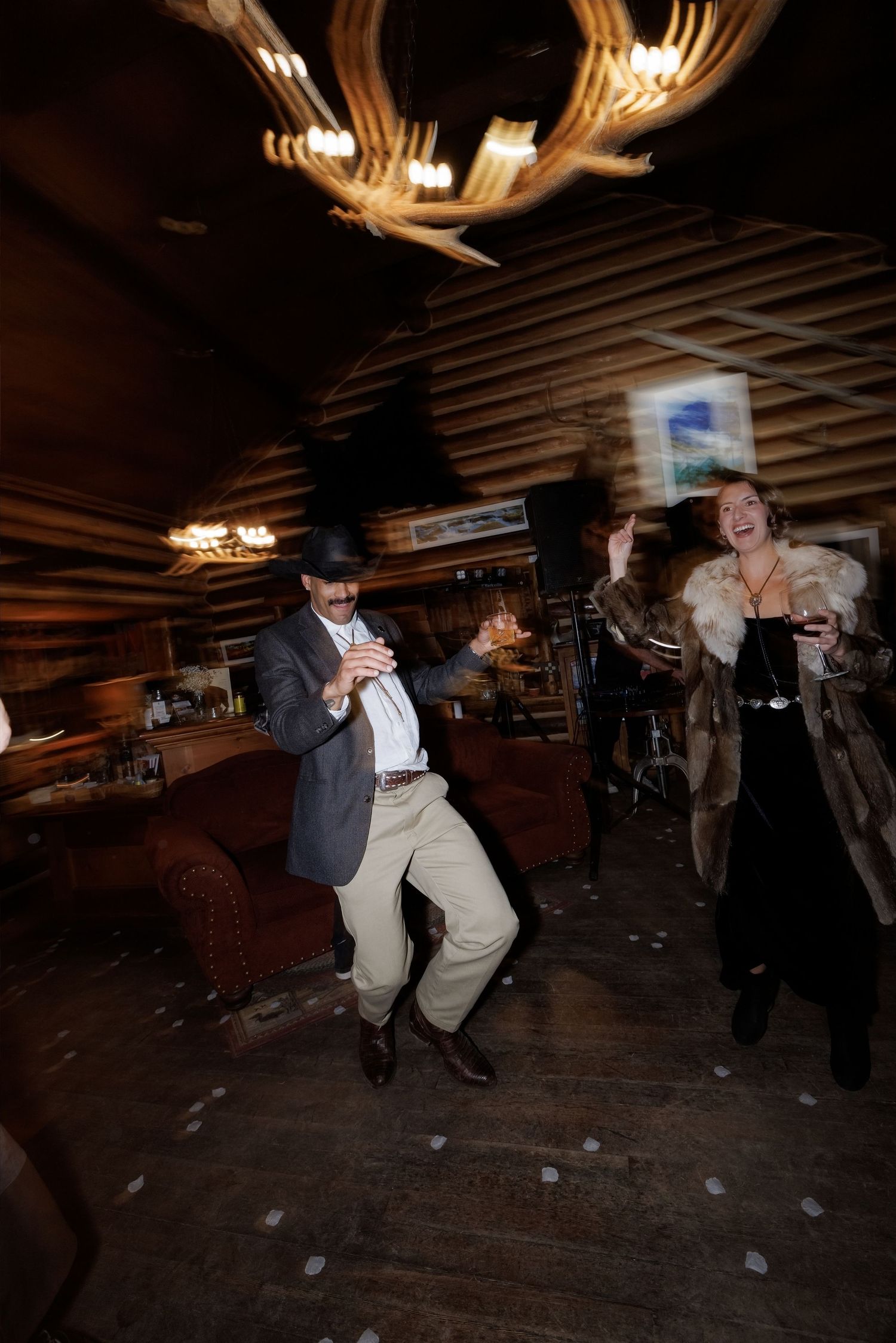 Guests dancing energetically in rustic log cabin wedding reception under antler chandelier with motion blur effect.