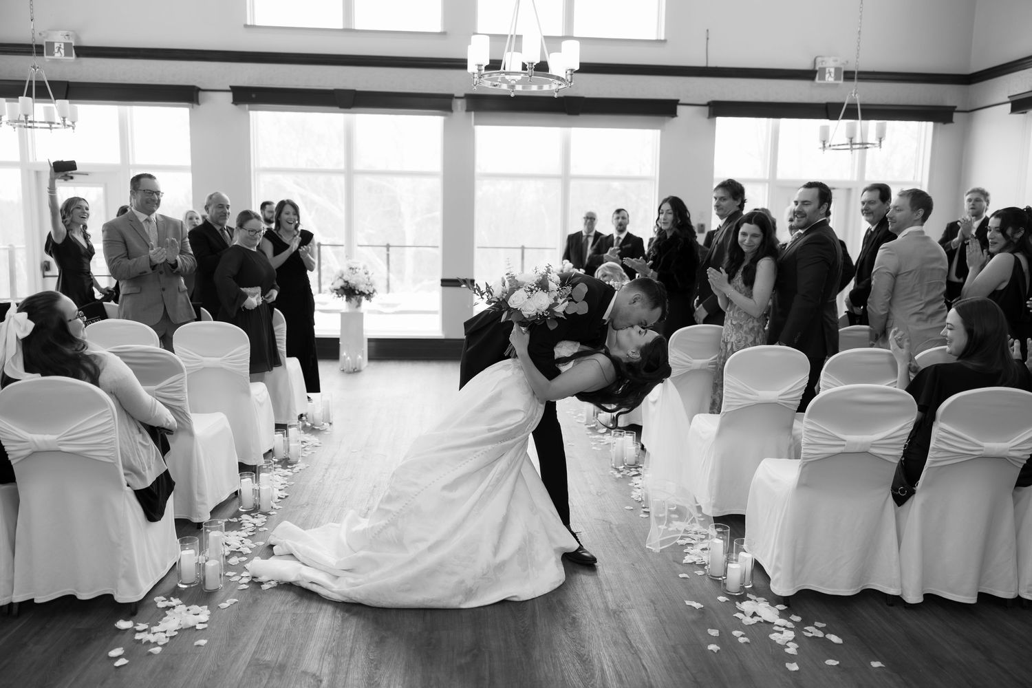 Black and white photo of bride and groom sharing a romantic dip kiss at wedding ceremony aisle.