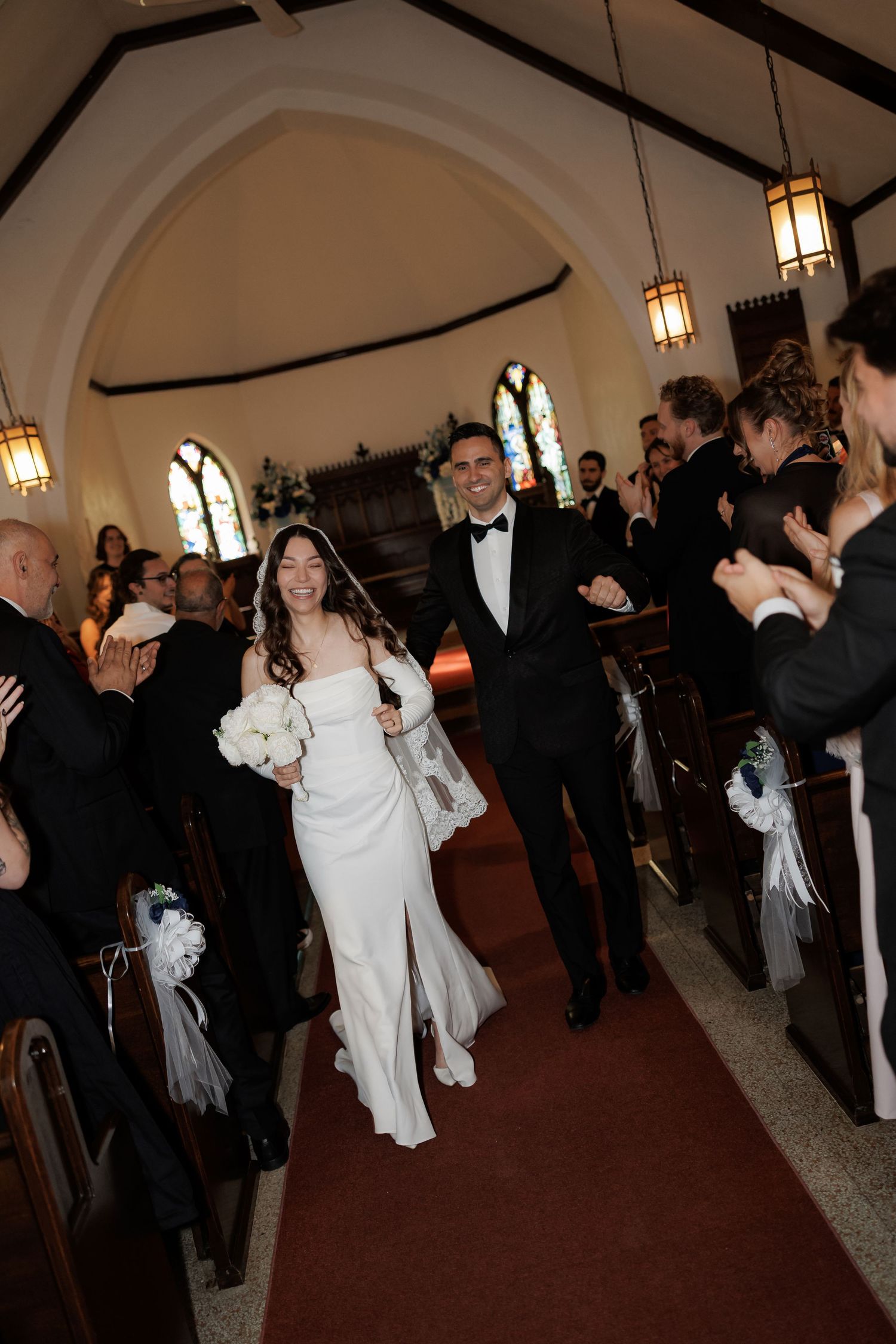 Wedding reception guests dancing and celebrating in an elegant ballroom with arched ceilings and hanging lights.