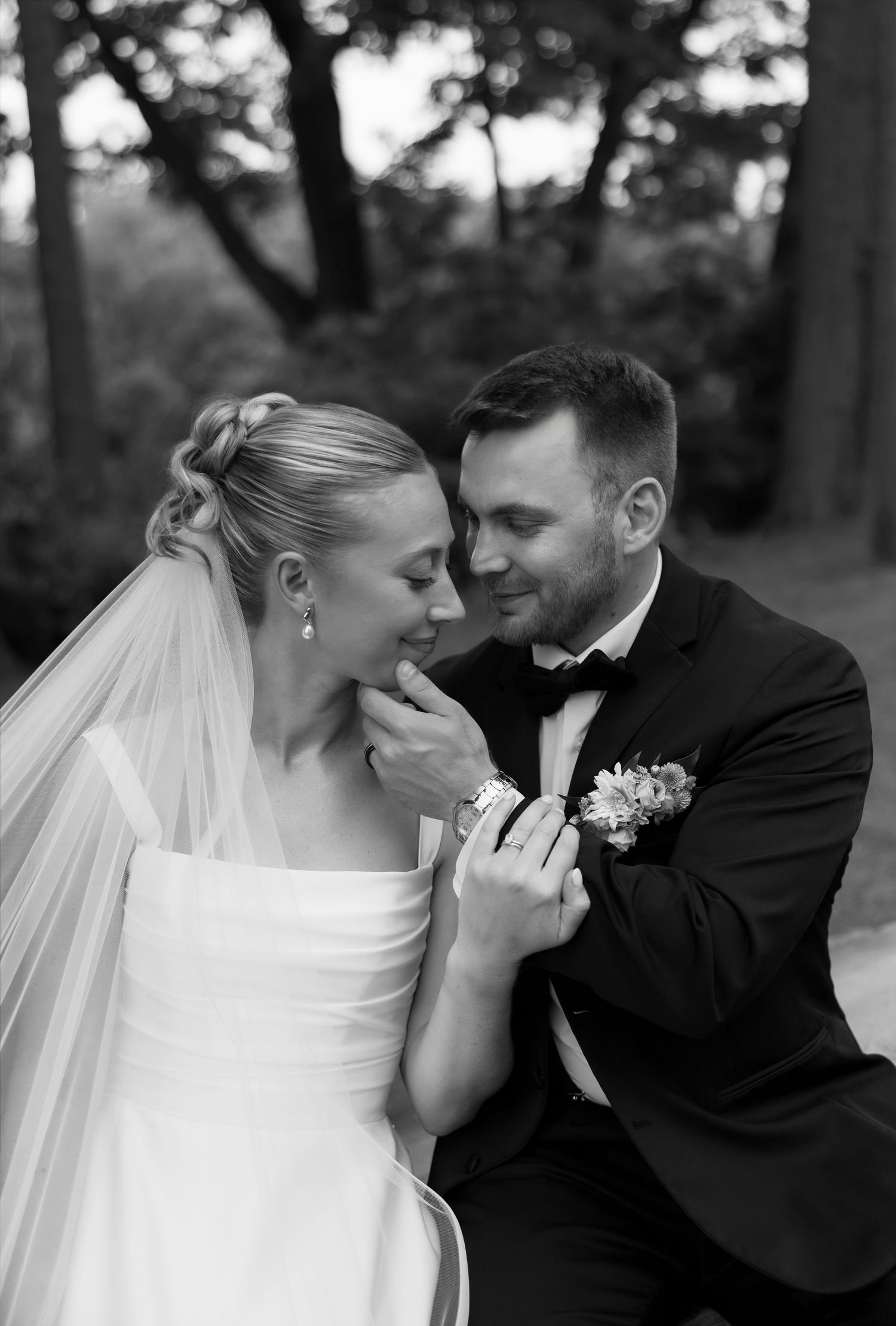 Black and white portrait of groom gently holding brides chin as she smiles in a garden setting.