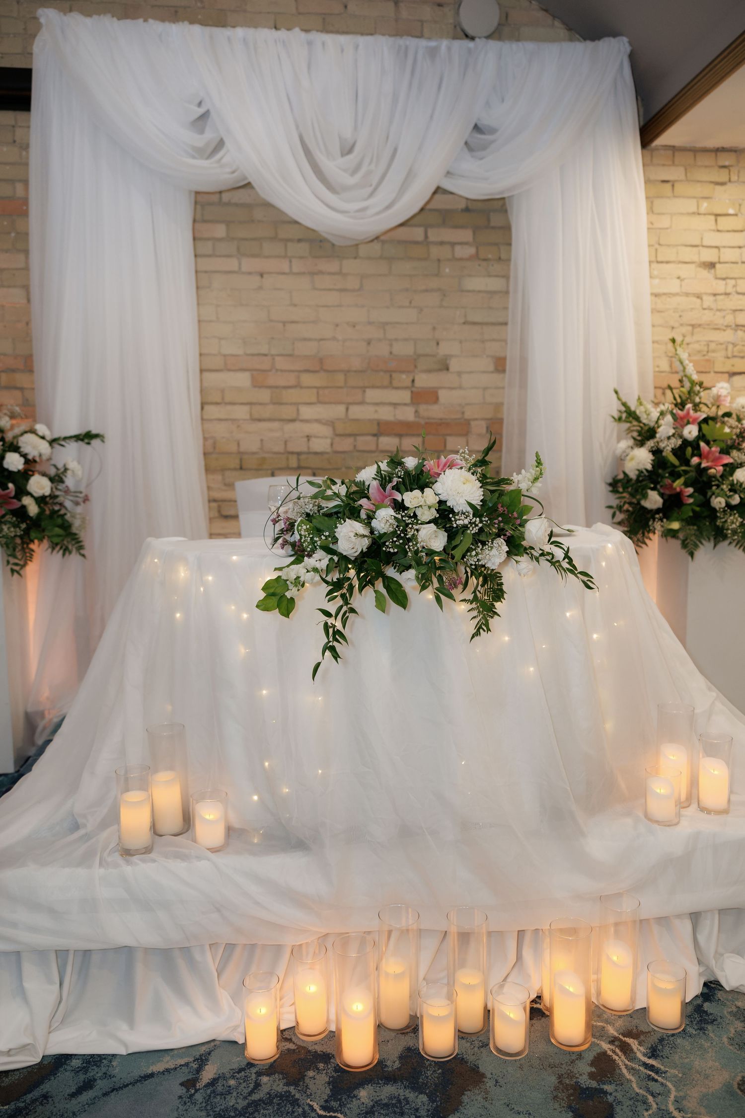 Elegant white draped backdrop with twinkling lights and floral arrangements for a wedding reception.