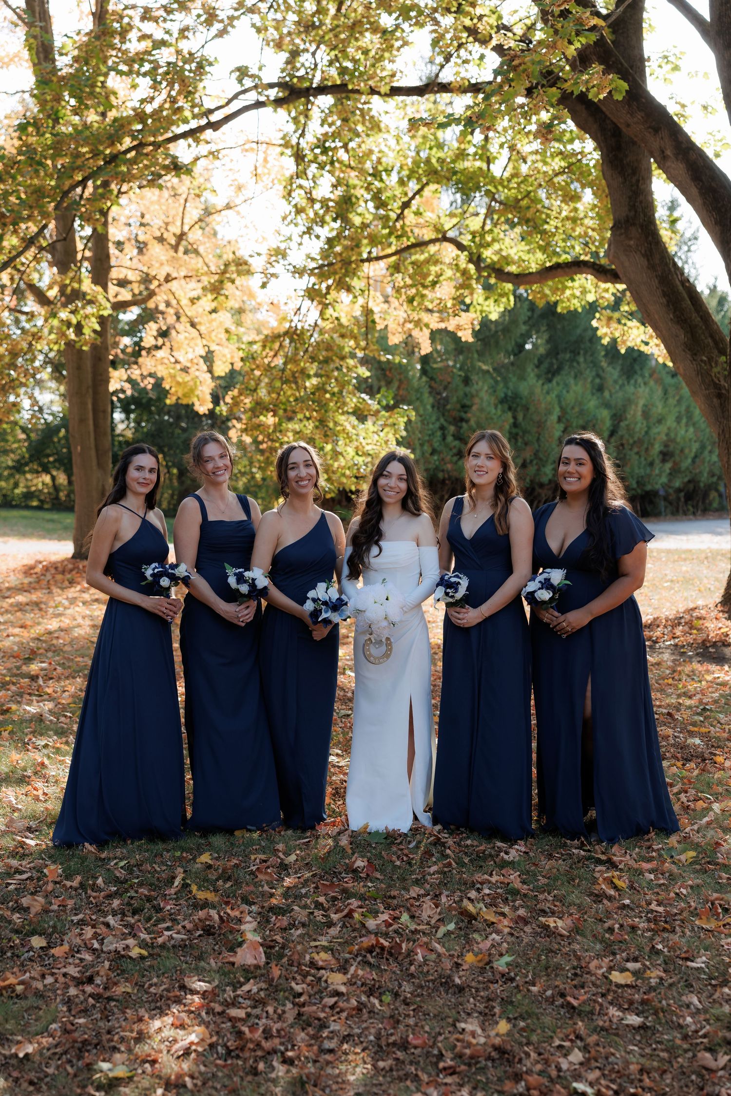 Bridesmaids in navy blue dresses stand with bride in white dress among fall foliage.