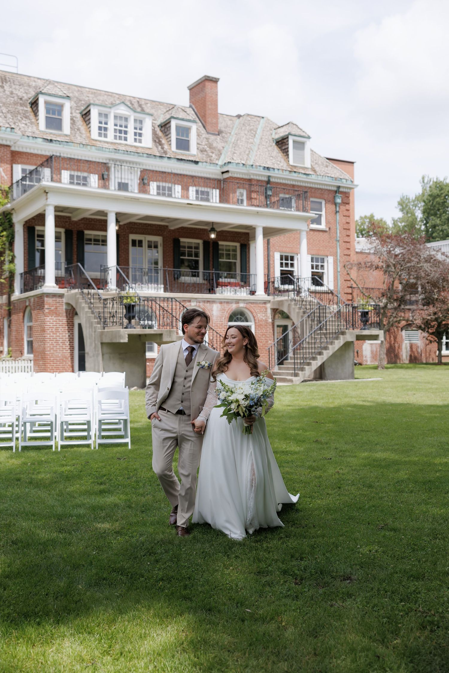 Bride and groom walk hand in hand on green lawn before elegant brick manor with white chairs set for outdoor wedding ceremony.