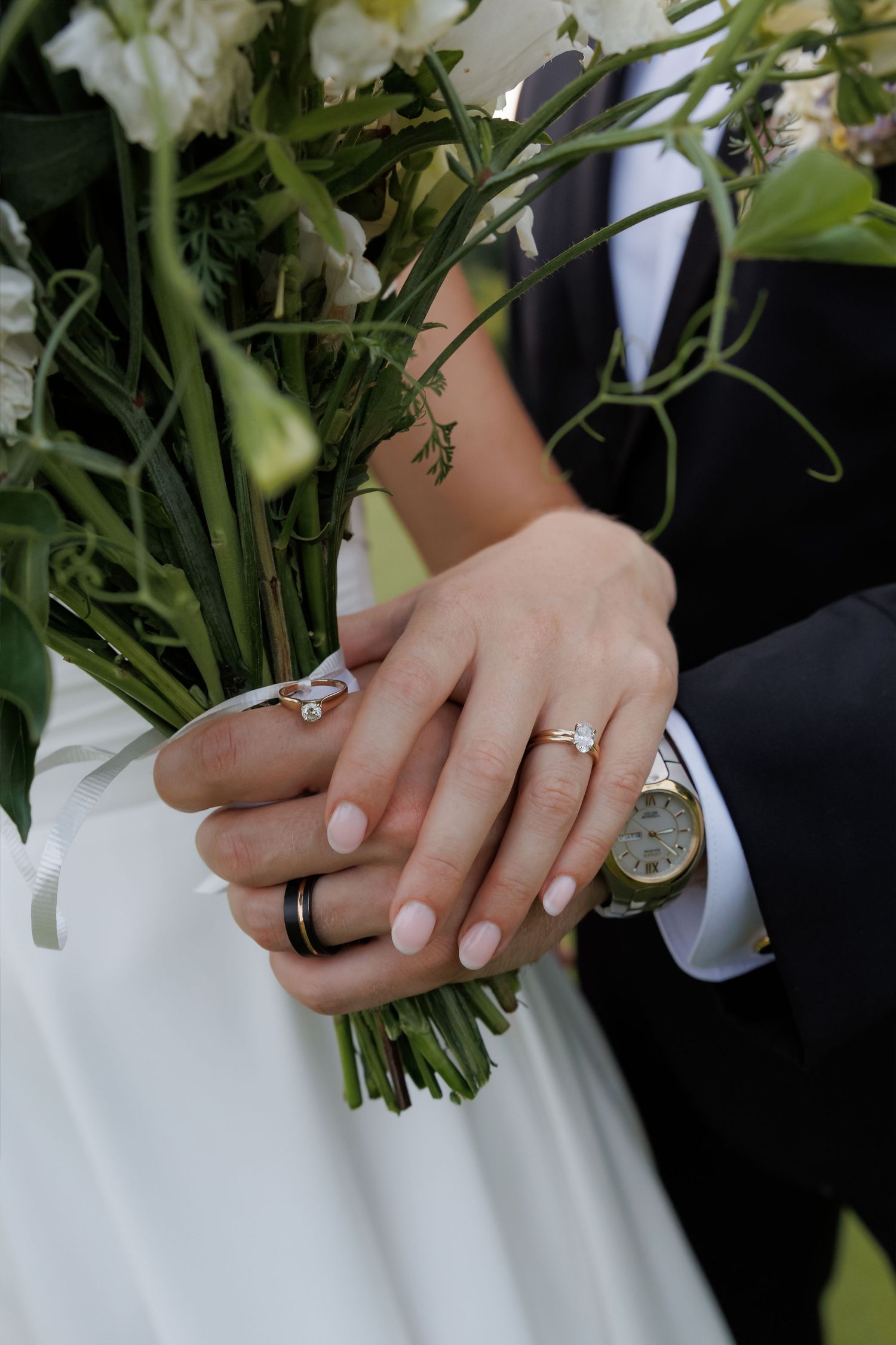 Close-up of newlyweds hands showing wedding rings while holding a white floral bridal bouquet.