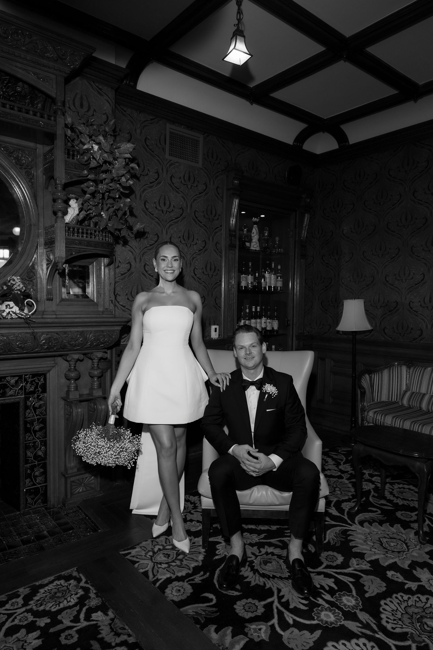 Bride in short white strapless dress and groom in tuxedo pose in elegant vintage room.