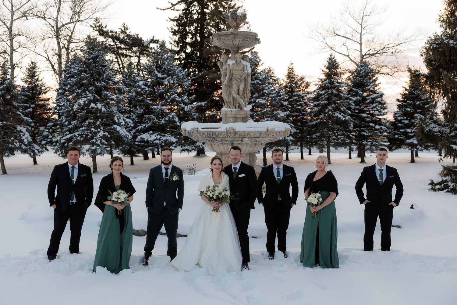 Winter wedding party poses in snow before fountain, bride in white, bridesmaids in green gowns, groomsmen in black suits.