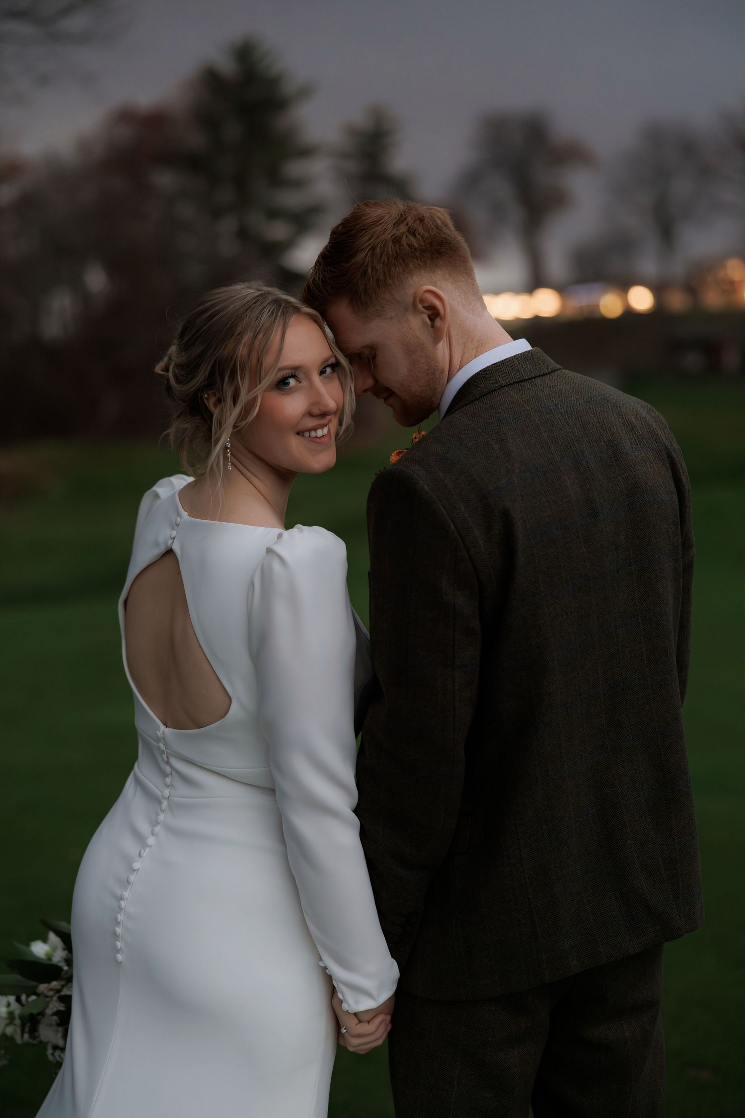 Bride in elegant white long sleeve dress shares intimate moments with groom in brown suit during outdoor wedding photoshoot.