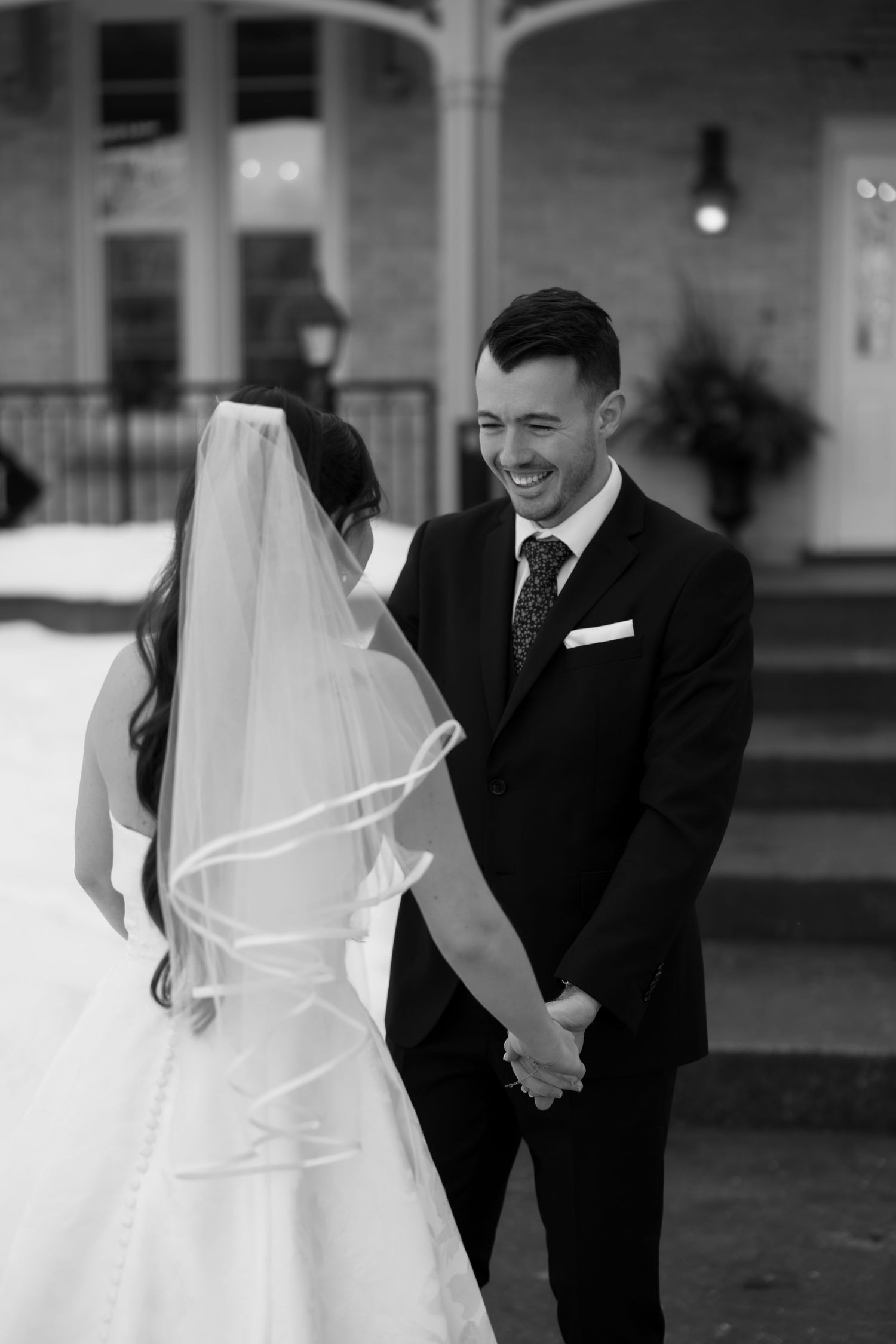 Black and white photo of a smiling groom in a suit holding hands with a bride in a wedding dress and veil outdoors.