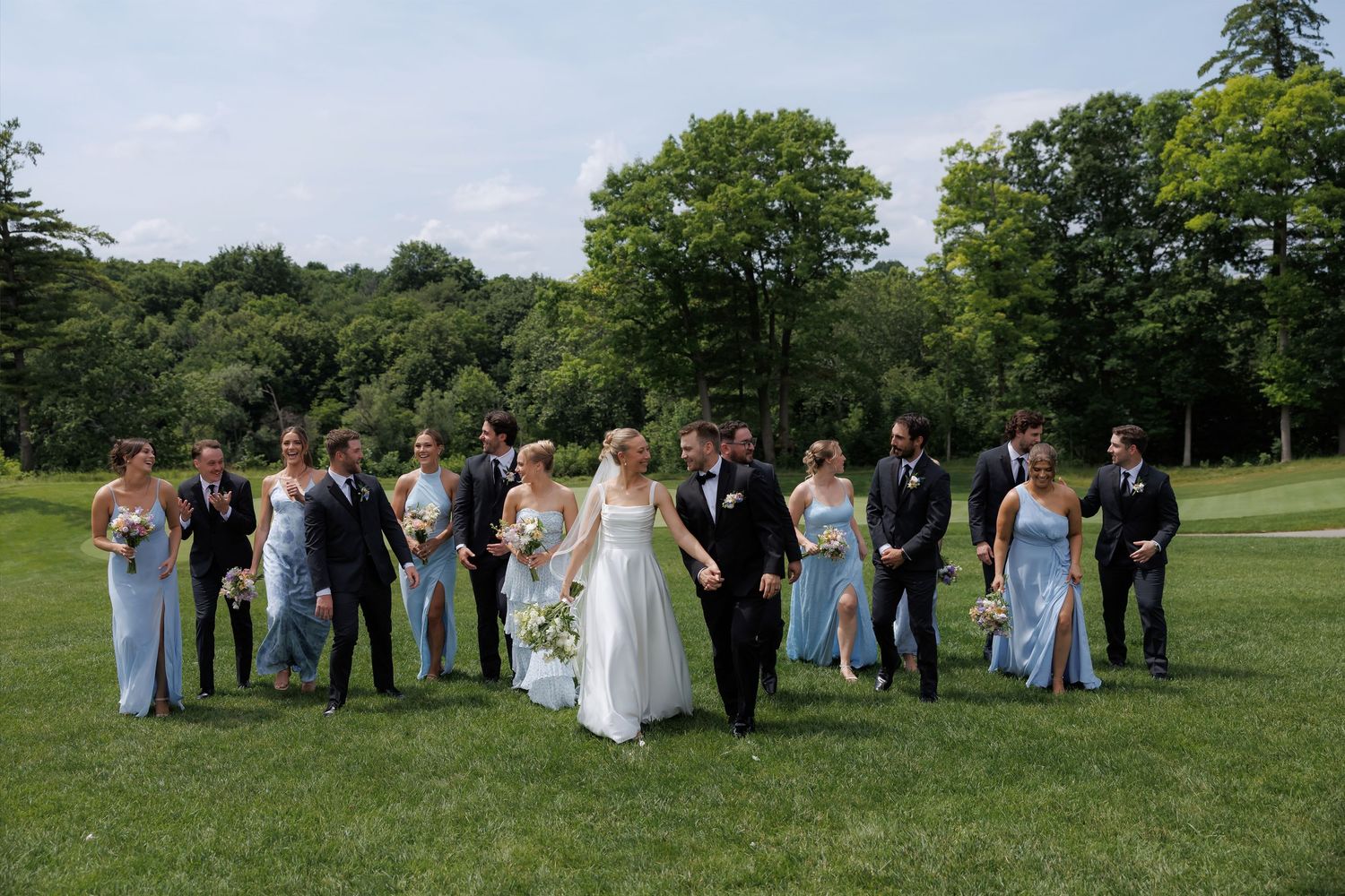 Wedding party walks outdoors on lush green lawn, bride in white gown with bridesmaids in blue dresses and groomsmen in dark suits.