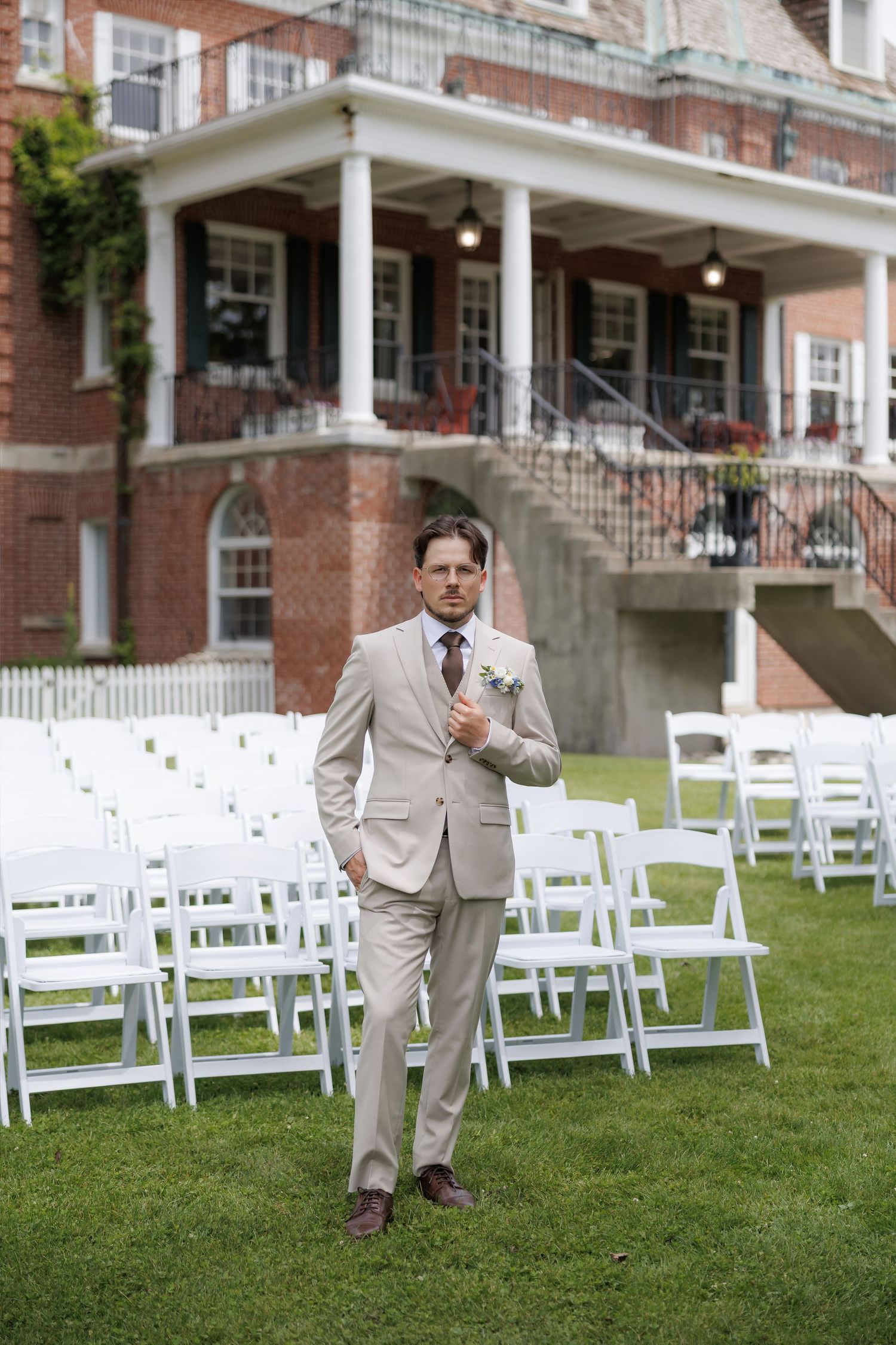 Groom in beige suit stands confidently on lawn before outdoor wedding ceremony with white chairs and brick venue.