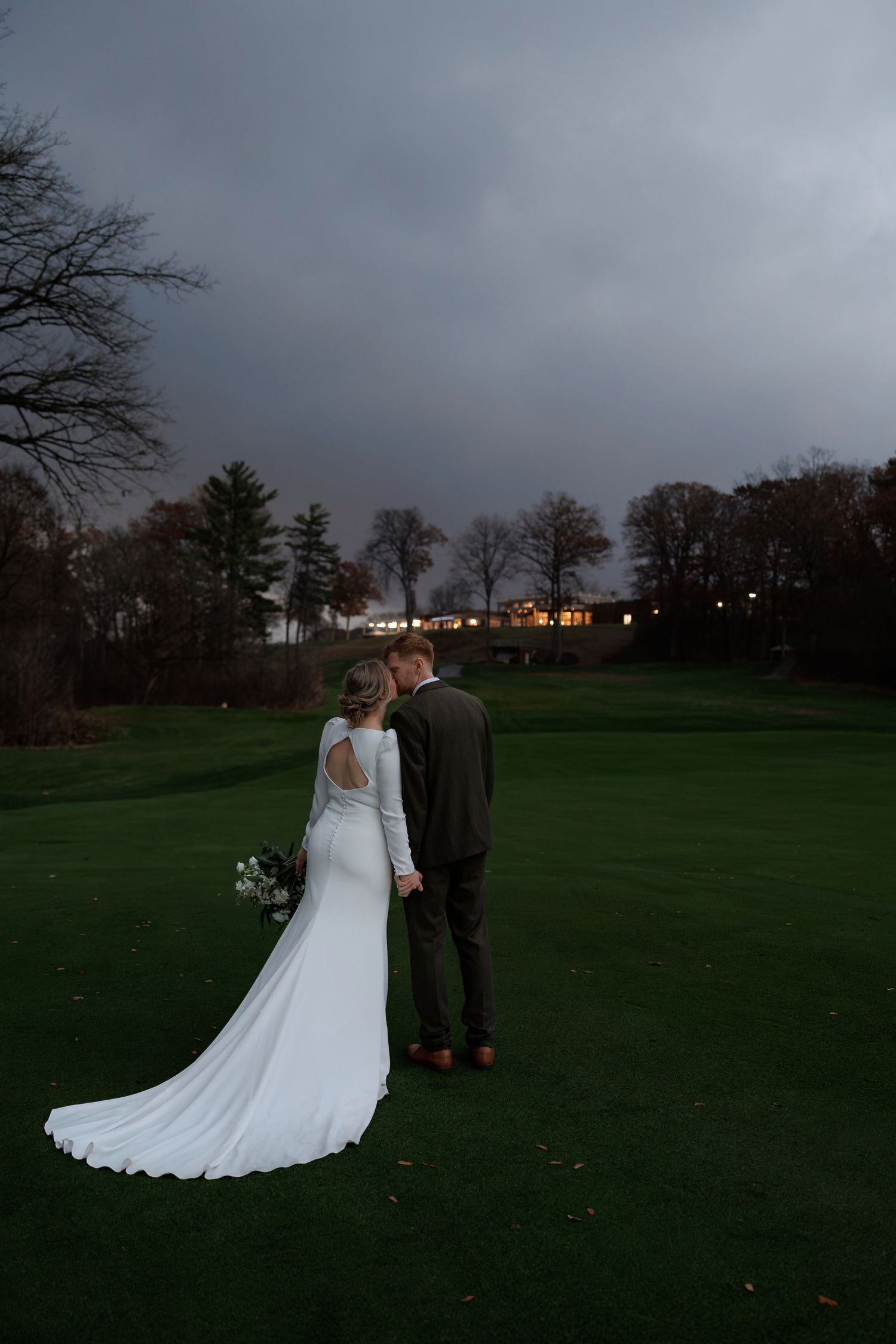 Couple in wedding attire stands on a dark golf course with city lights glowing in the distance.