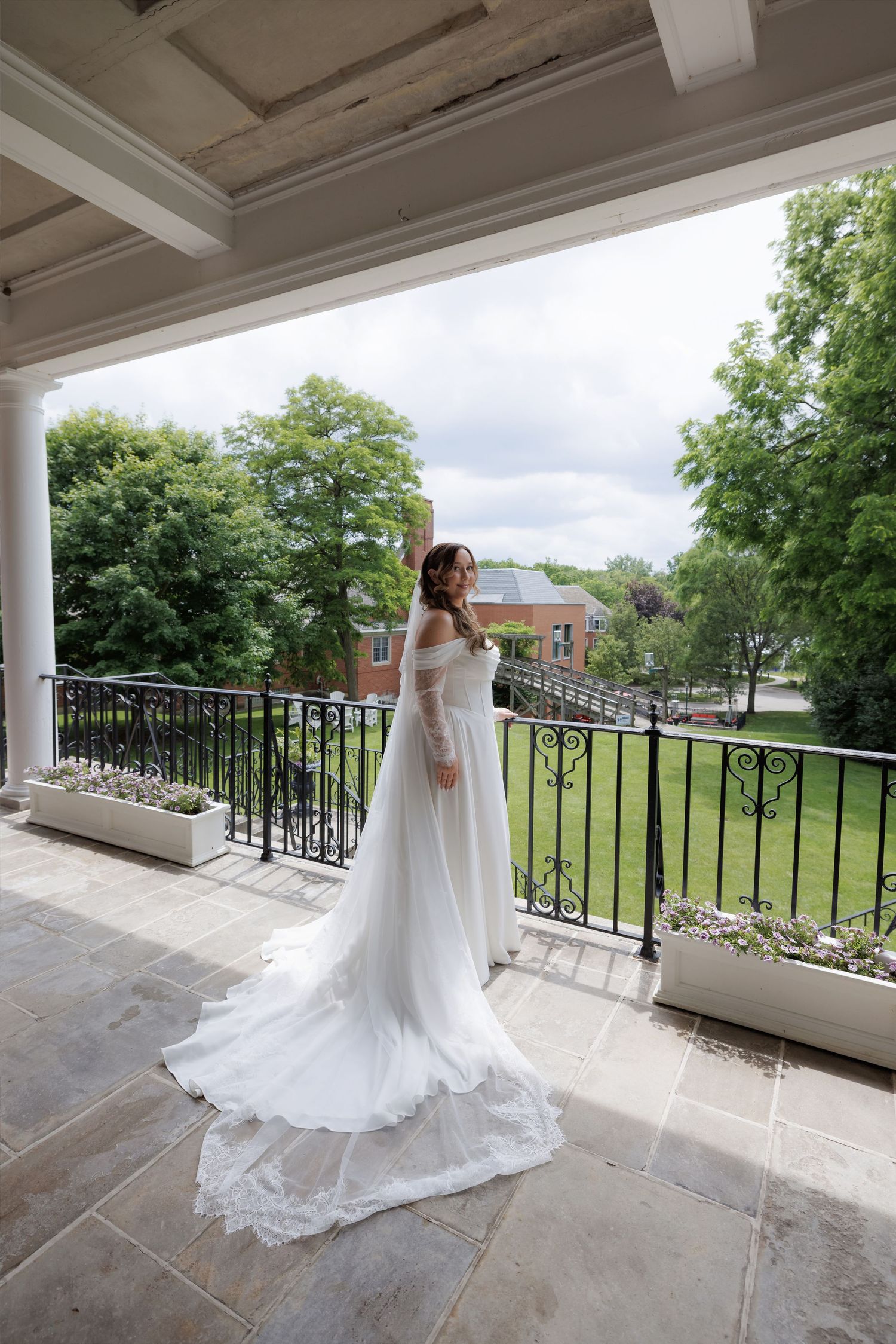 Bride in elegant white off-shoulder gown with long train stands on ornate balcony overlooking lush garden.
