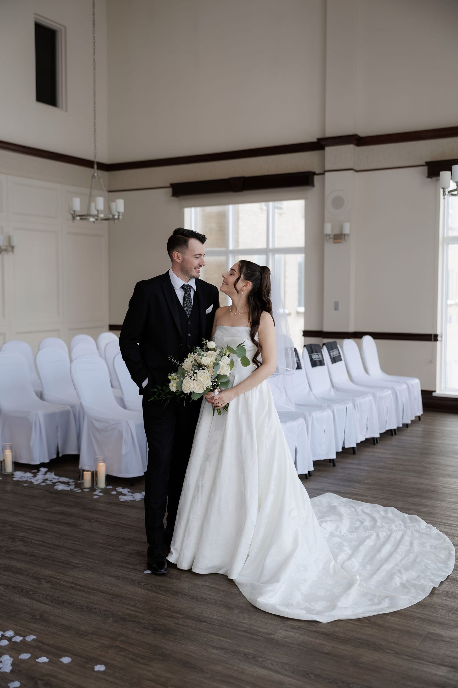 Bride and groom share a romantic moment in elegant white ceremony hall with candles and draped chairs.
