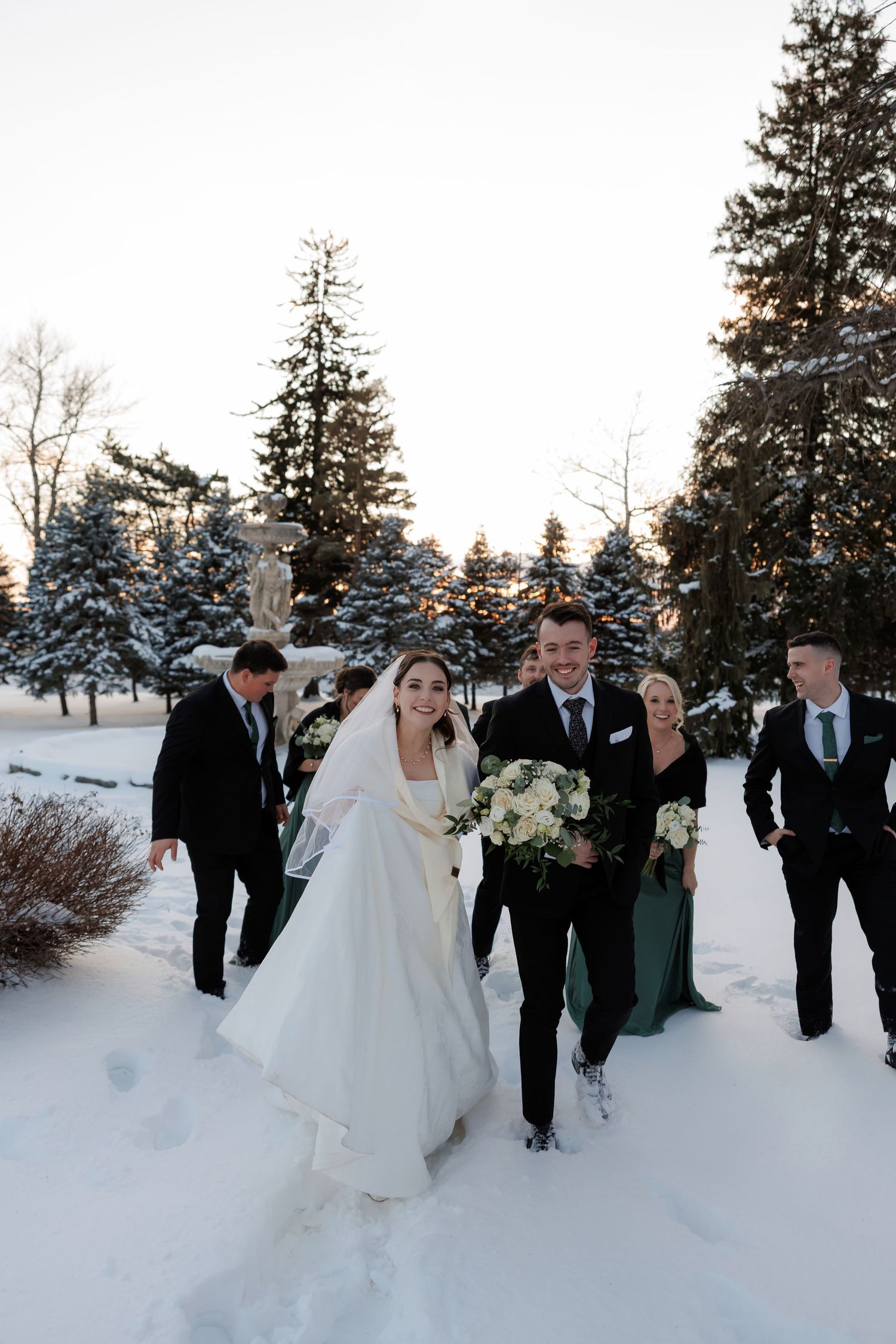 Wedding party walks through snowy winter landscape, bride in white cape holding bouquet with groomsmen and bridesmaids.
