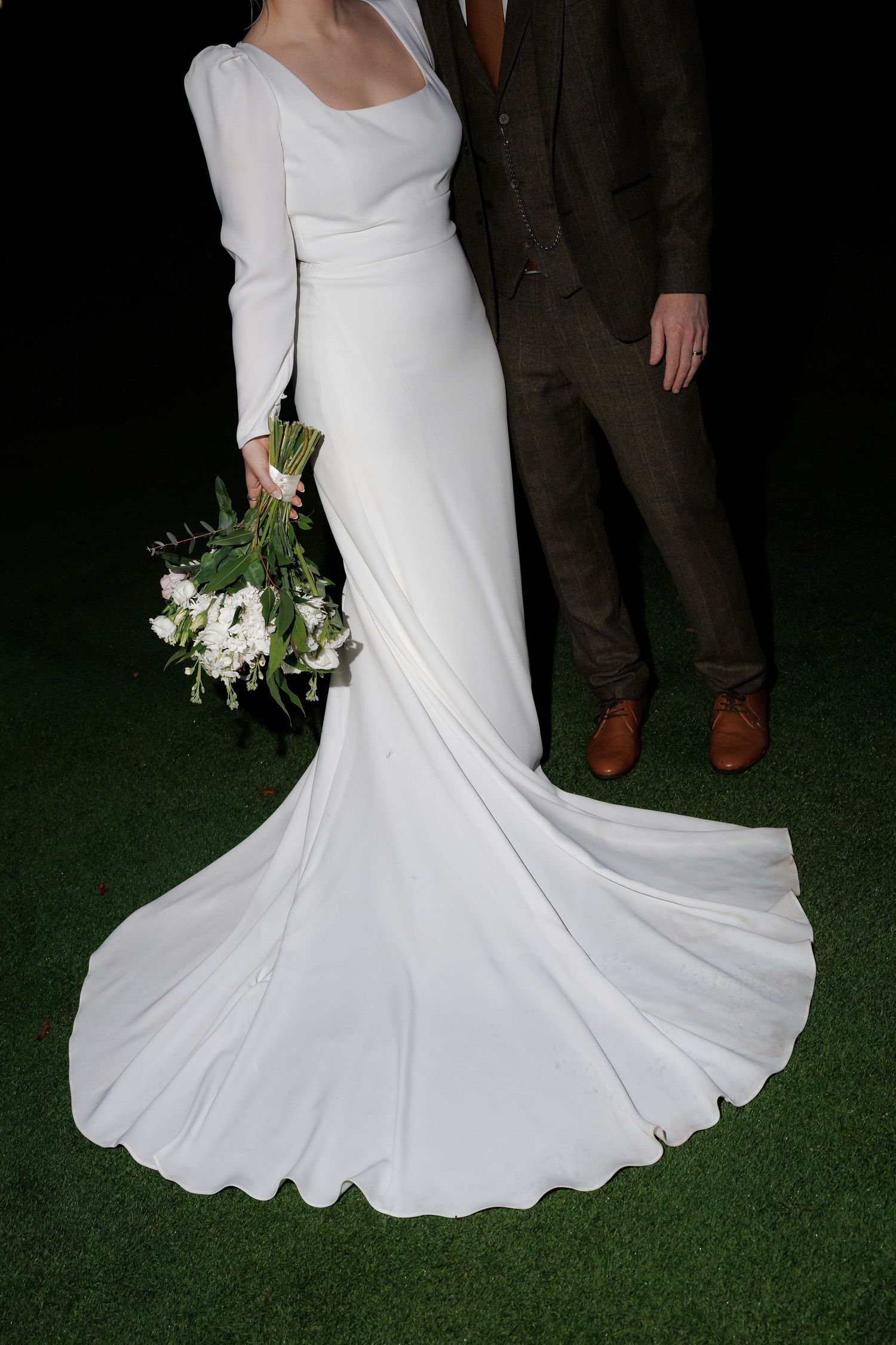 Close-up details of a flowing white wedding dress train against green grass with a delicate white floral bouquet.