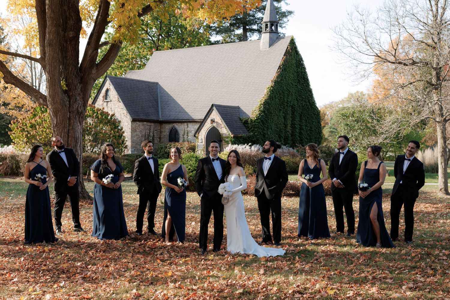 Wedding party members spread out in a line across fallen autumn leaves near a church.
