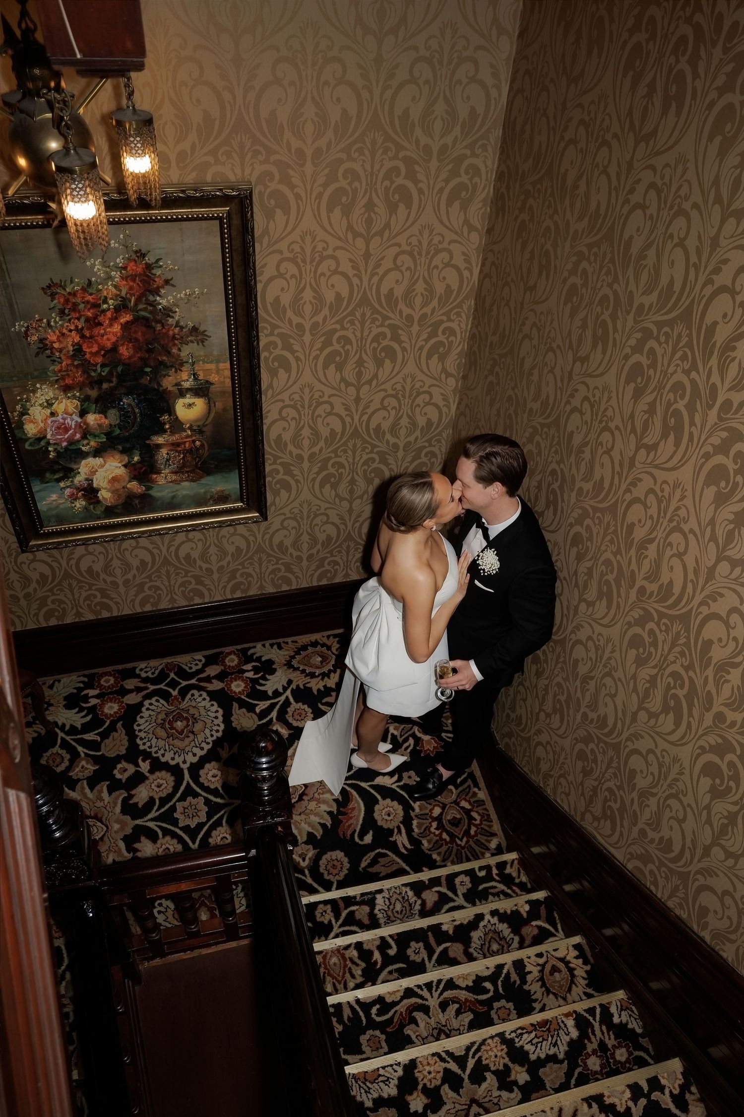 Bride and groom share a romantic kiss on an ornate staircase with floral wallpaper and a vintage painting nearby.
