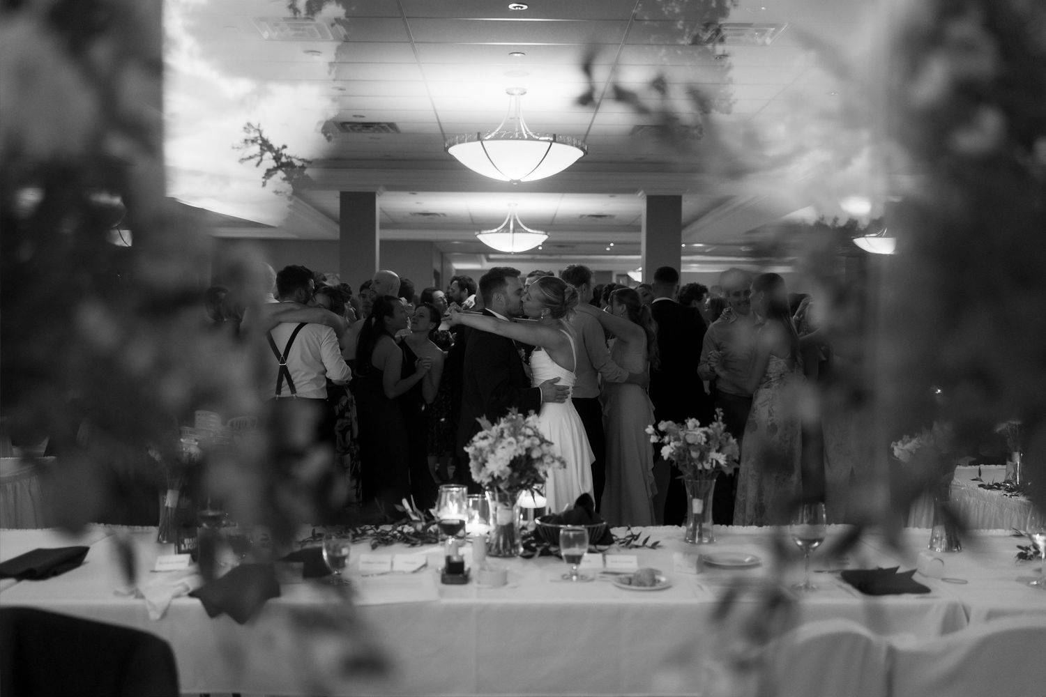 Black and white wedding reception photo showing guests dancing and embracing in elegant ballroom with floral centerpieces.