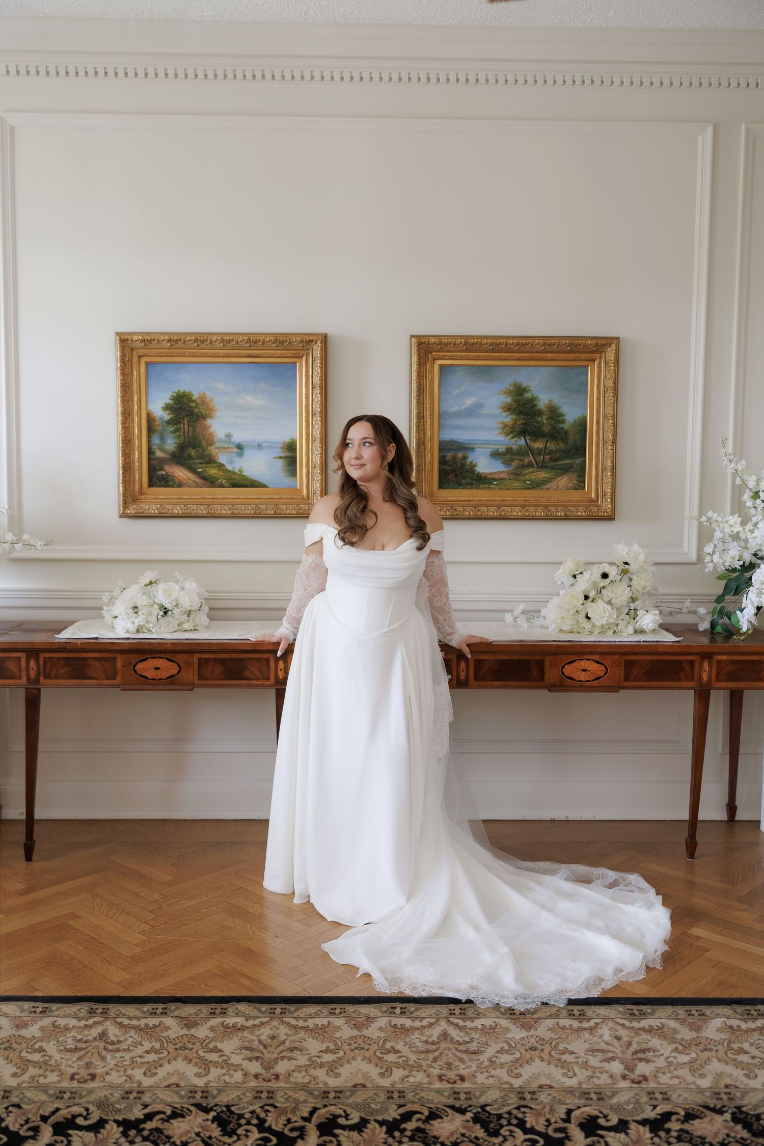 Bride in a flowing white off-shoulder gown with lace sleeves stands before gold-framed paintings in an elegant room.