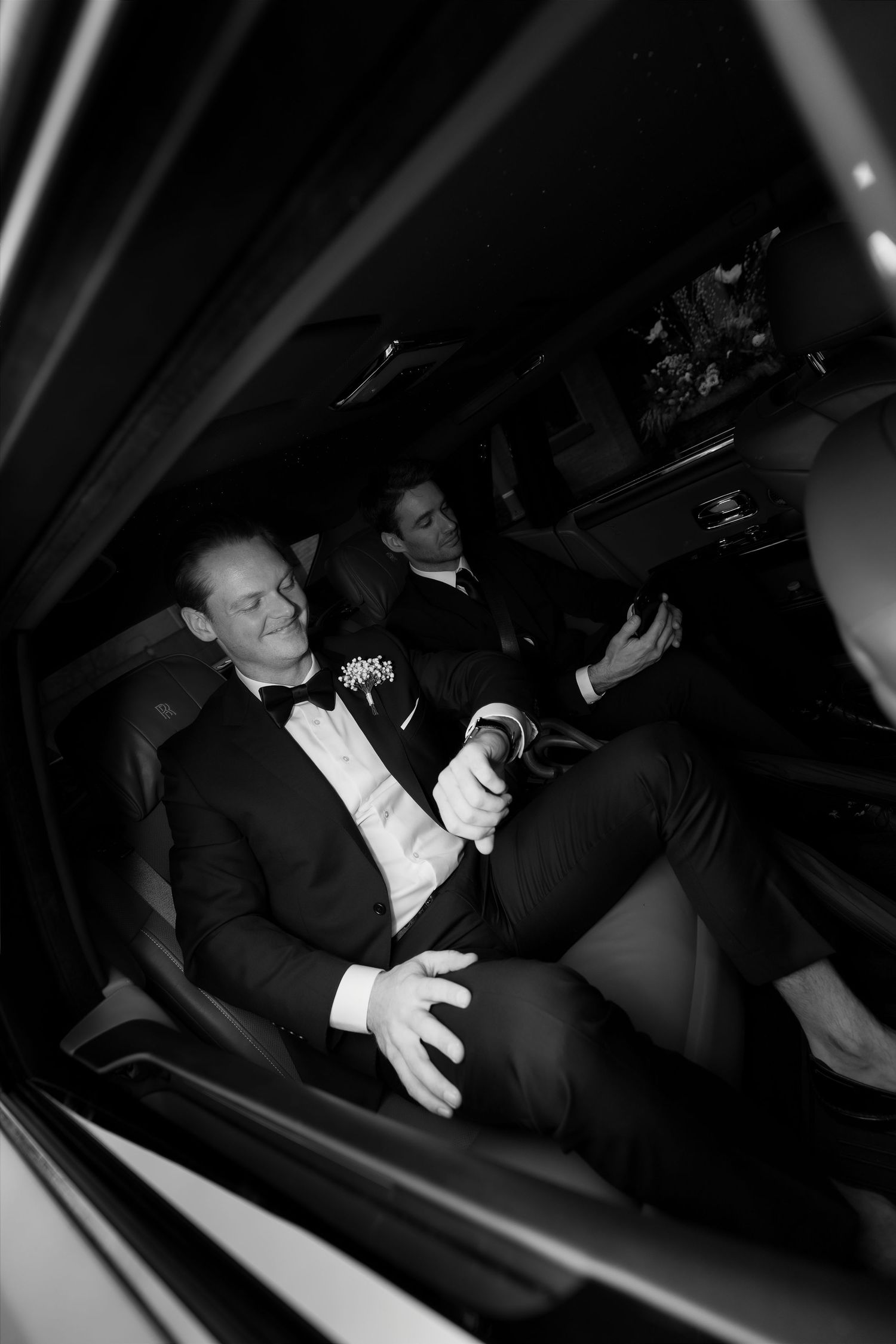Two grooms in tuxedos relax inside a limousine, one wearing a boutonniere, in a black and white wedding photo.