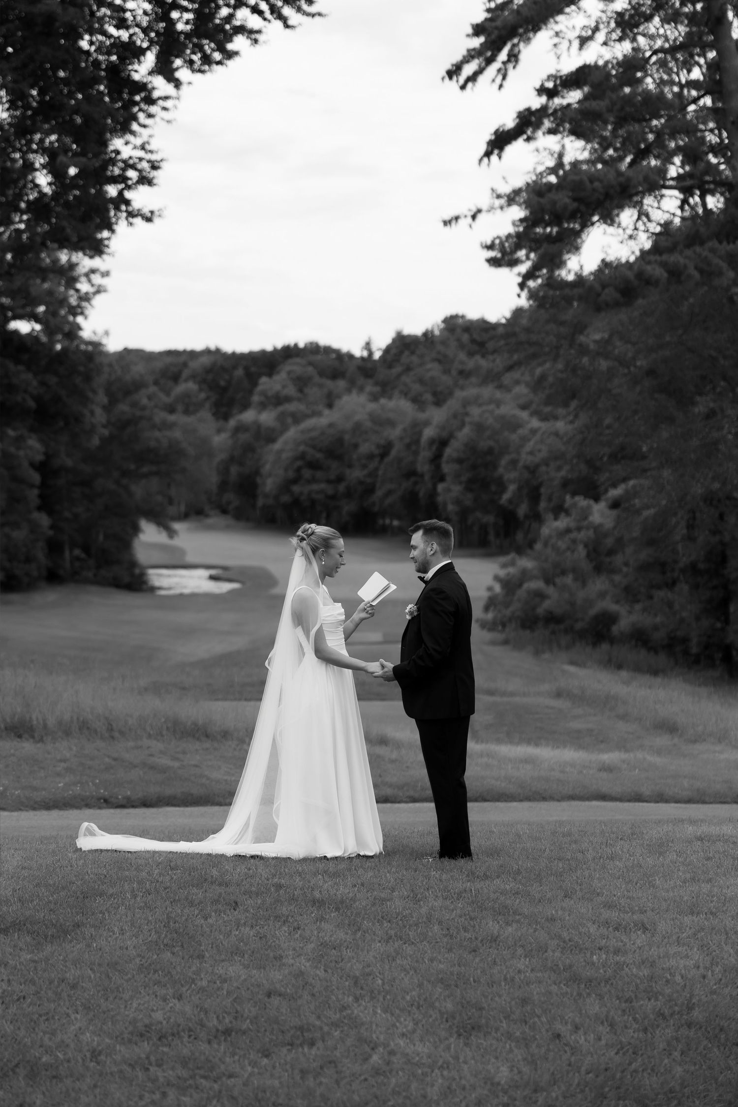 Black and white photo of bride and groom exchanging vows on a golf course surrounded by lush trees.