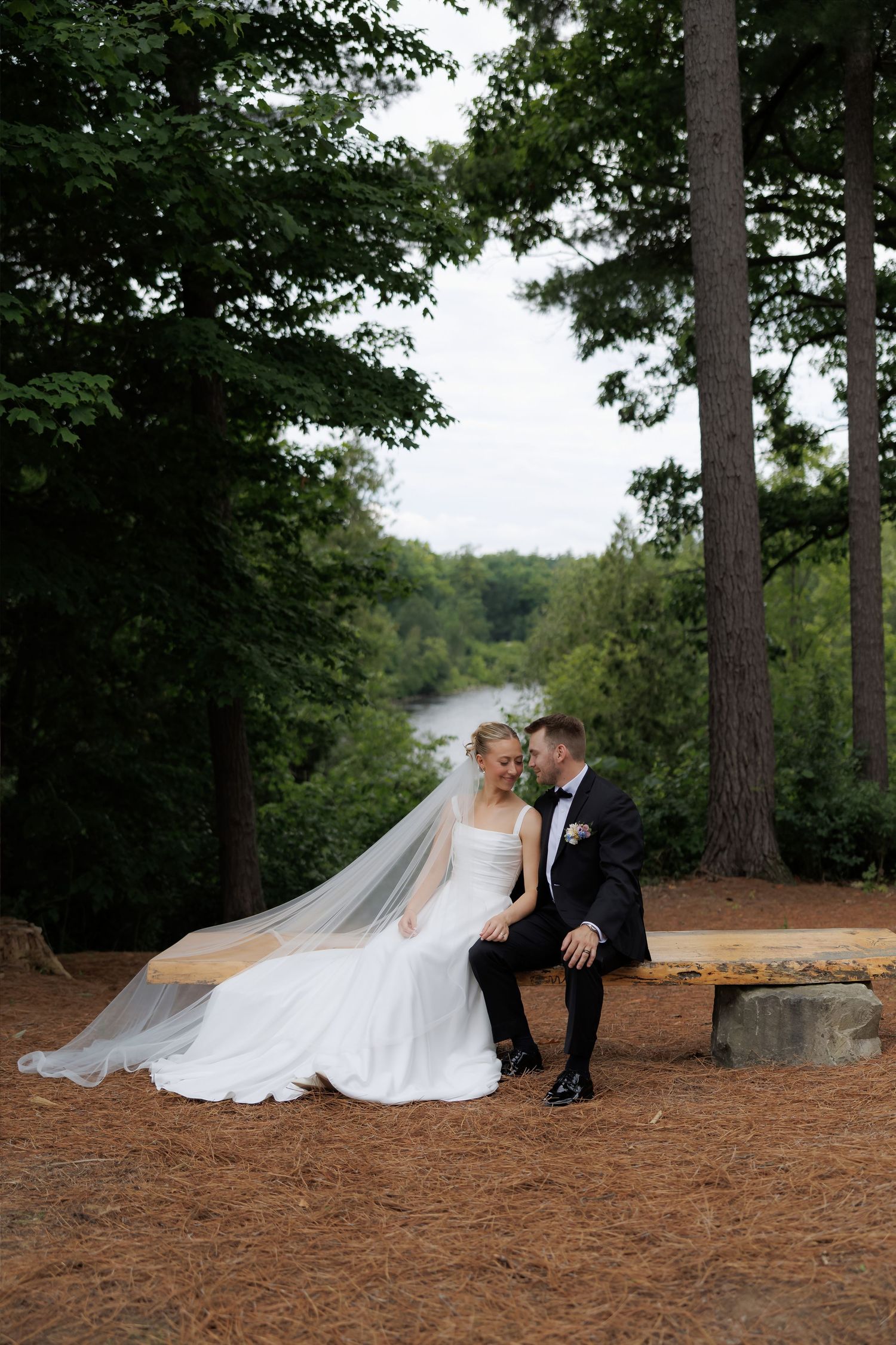 Bride and groom sit on a wooden bench surrounded by tall pines and a river view on their wedding day.