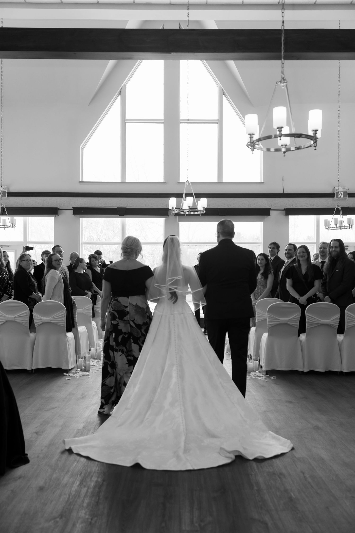 Bride walks down the aisle escorted by parents in elegant chapel with vaulted windows.