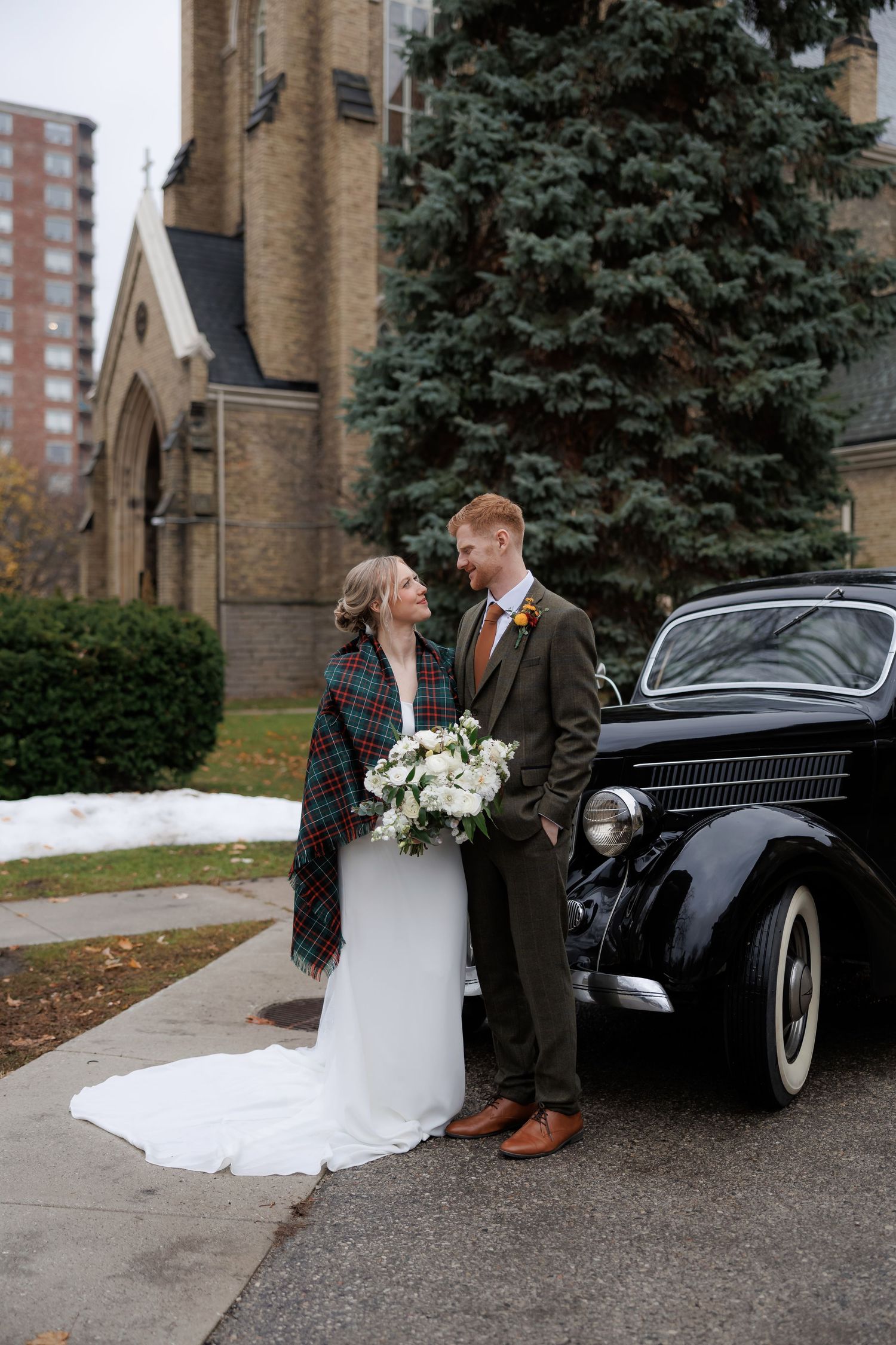 A couple poses with a vintage black car outside a historic church on their wedding day.