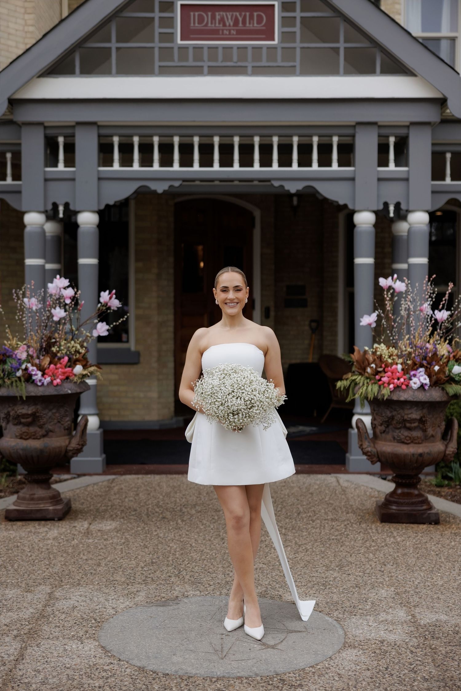 Smiling bride in short strapless white dress holds baby's breath bouquet outside Idlewyld Inn entrance.