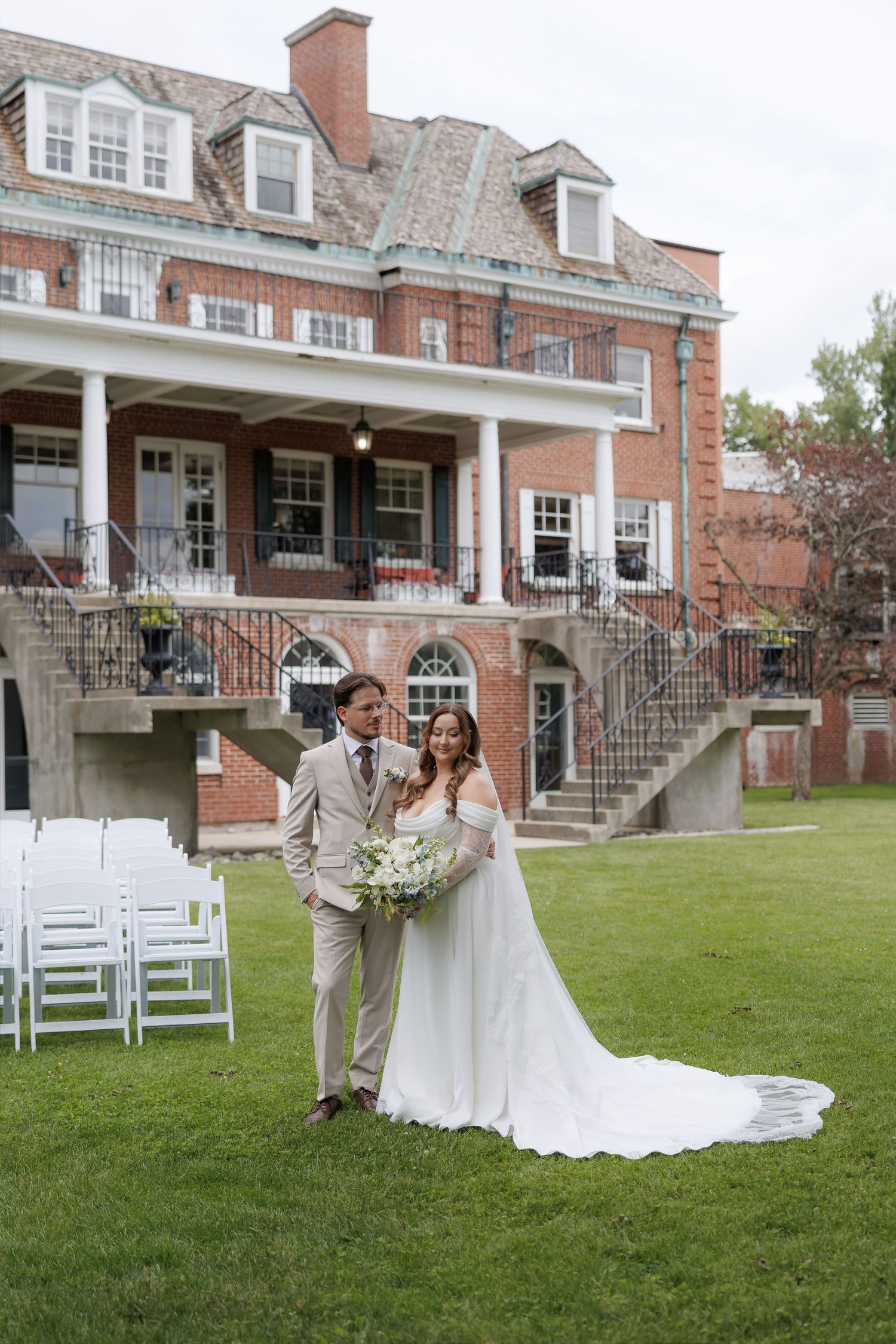 Bride and groom standing on green lawn before grand brick estate with white columns and stone staircase.