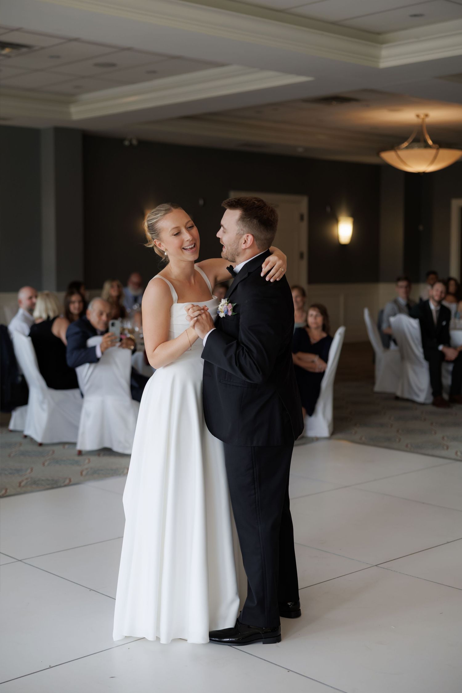 Bride and groom share a joyful first dance, smiling and embracing in an elegant wedding reception ballroom.