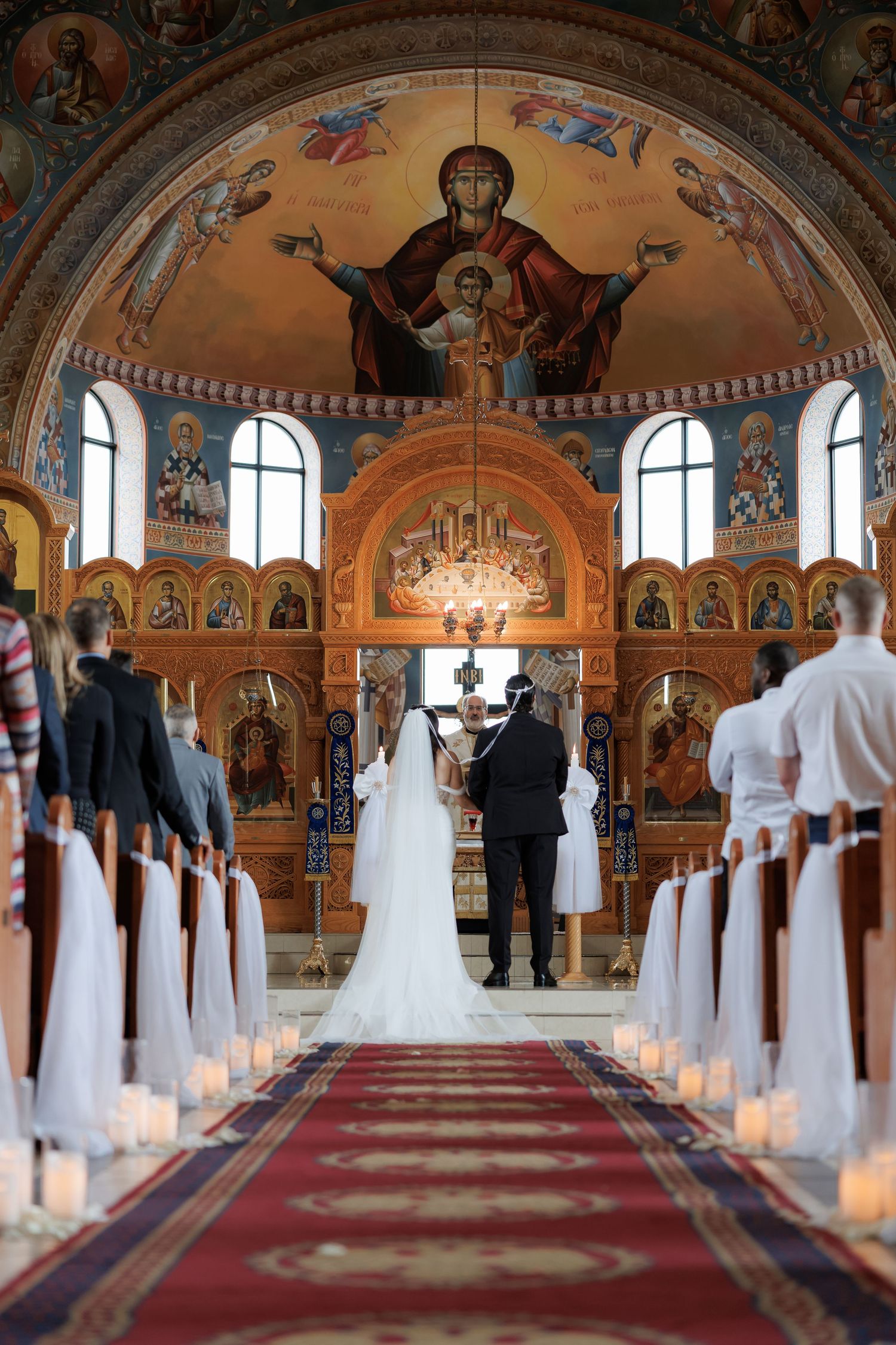 Wedding ceremony in Orthodox church with illuminated candles lining red carpet aisle.