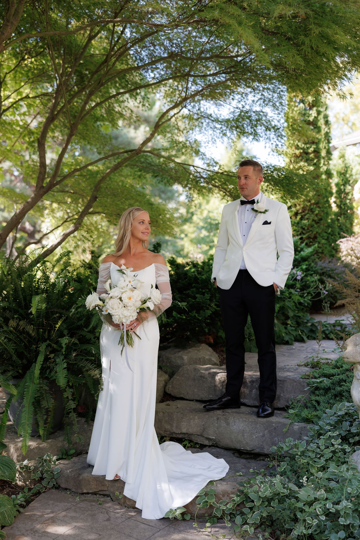 Wedding couple stands together on a garden path with beautiful foliage and dappled sunlight.