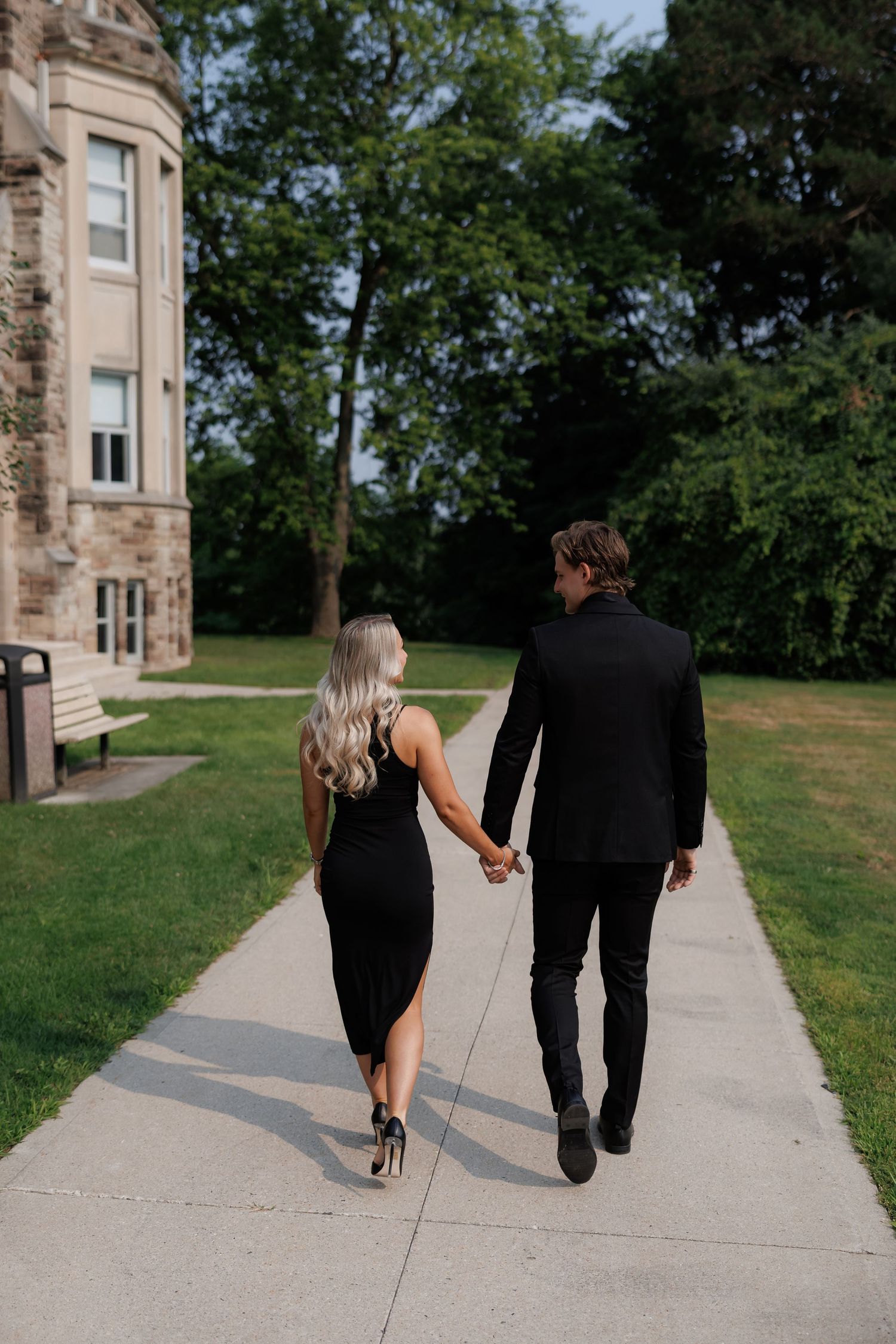 Couple in black formal wear walking hand in hand on a path near a stone building.
