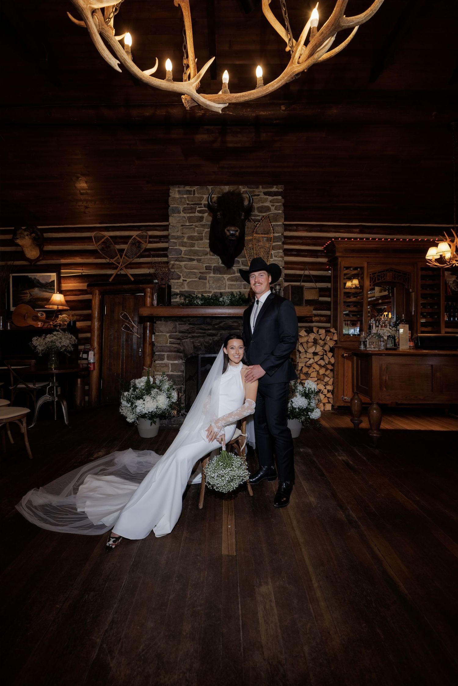 Bride and groom pose in rustic log cabin wedding venue with antler chandelier and stone fireplace.