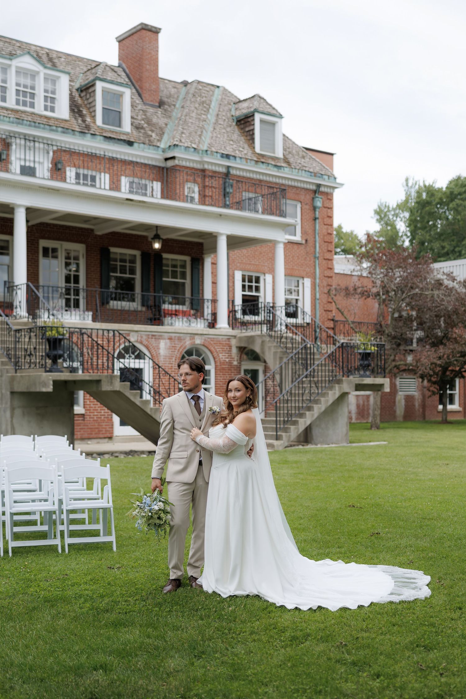 Bride and groom pose on green lawn before elegant brick mansion with white chairs set for outdoor wedding ceremony.