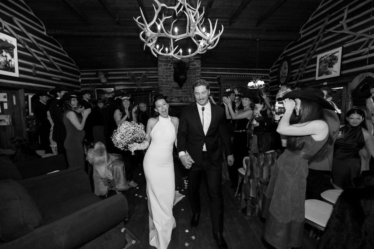 Black and white photo of bride and groom walking through cheering guests in rustic log cabin venue.