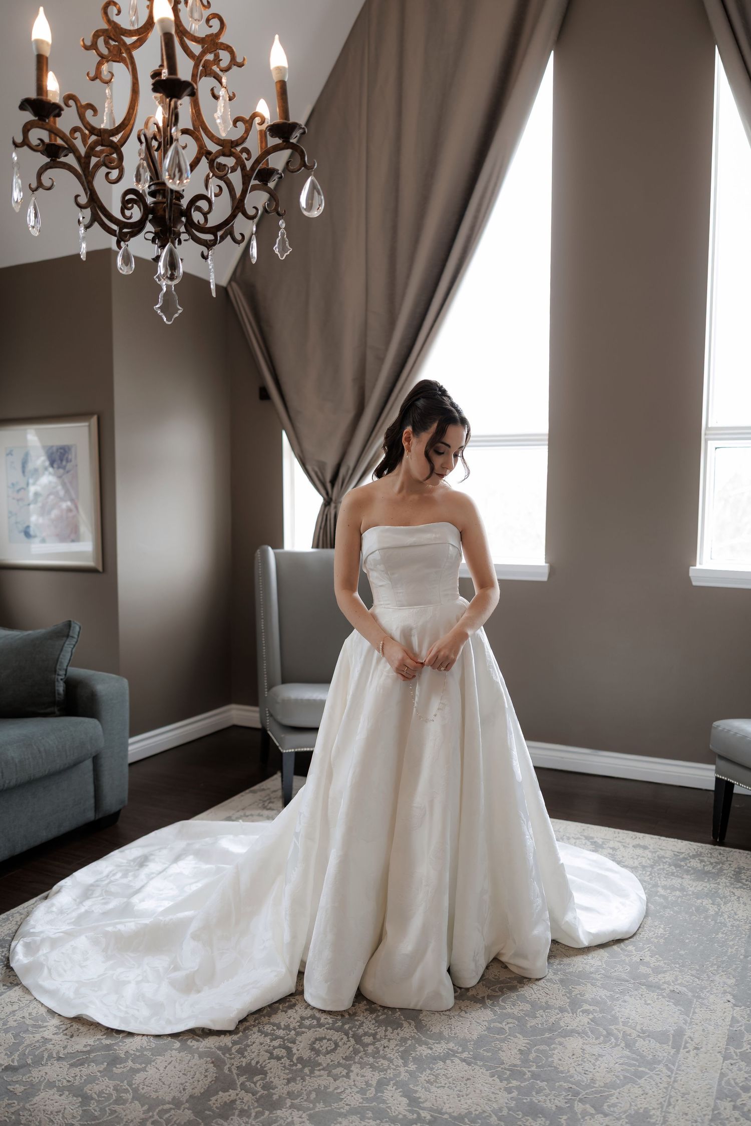 Bride in strapless ballgown wedding dress with long train standing in elegant room with crystal chandelier.