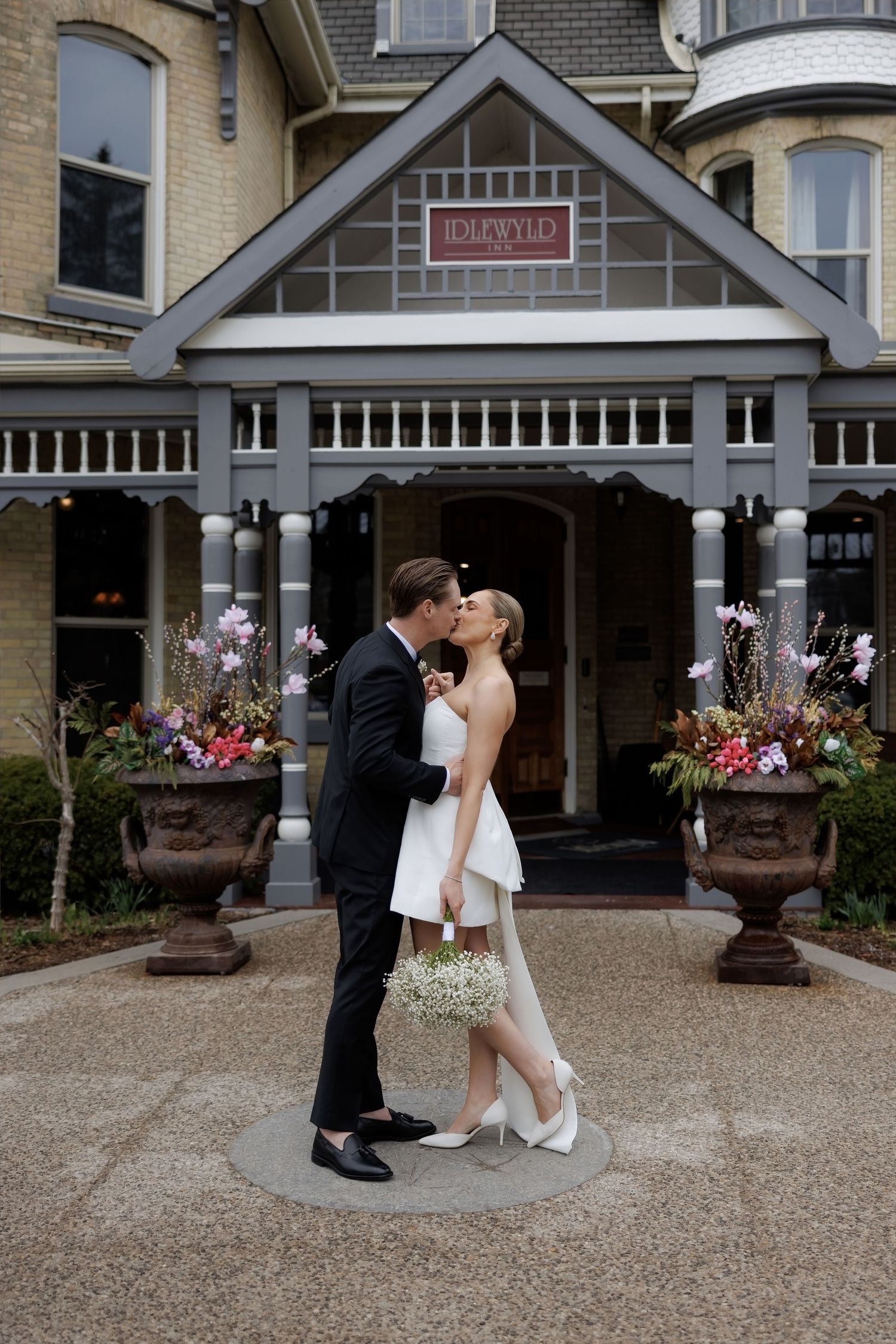 Bride and groom kissing in front of Idlewyld Inn entrance, flanked by floral urns, bride in short white dress.