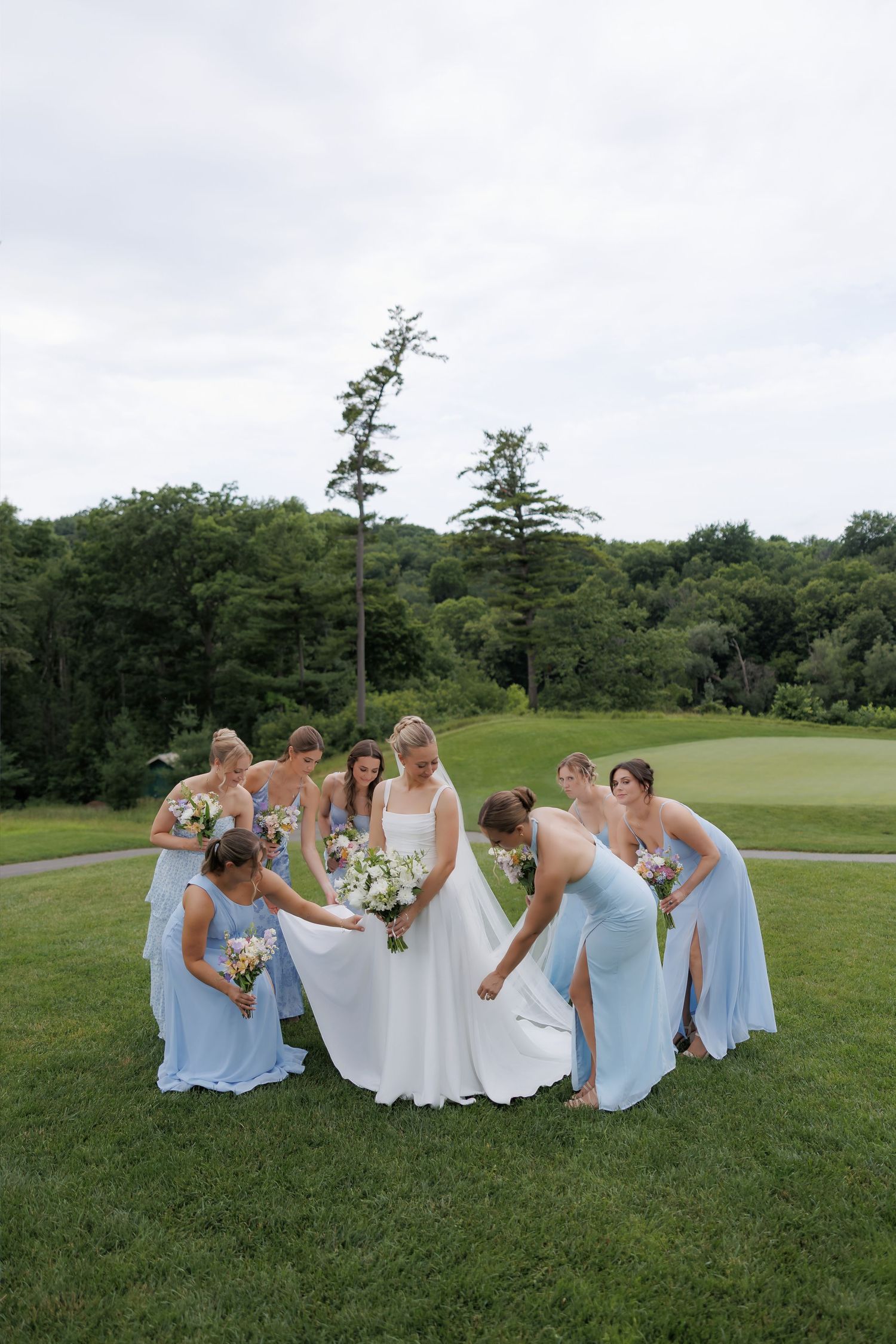 Bride in white dress surrounded by bridesmaids in light blue gowns arranging her train on a green lawn.