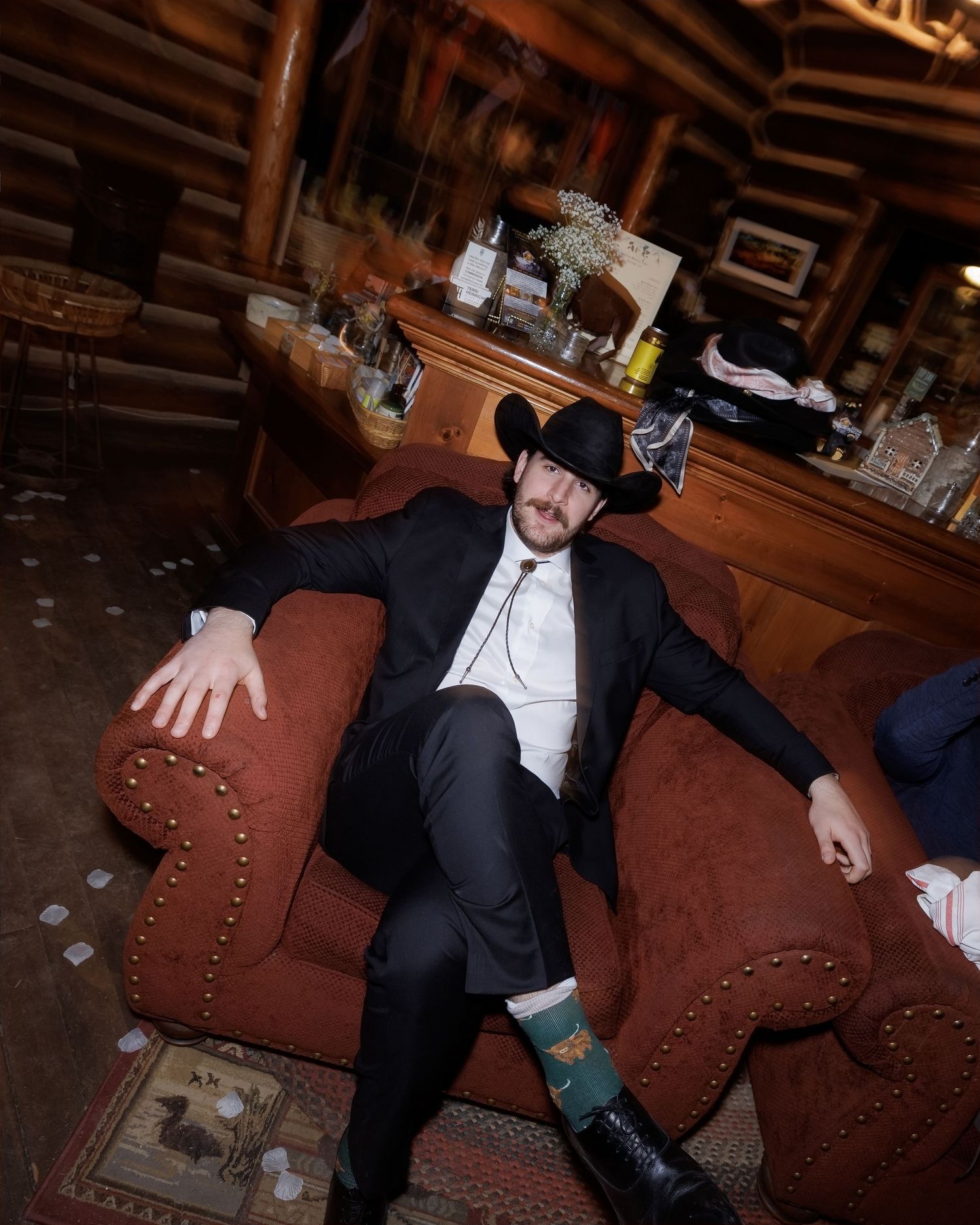 Groom in black suit and cowboy hat relaxes on red sofa at rustic log cabin wedding reception.