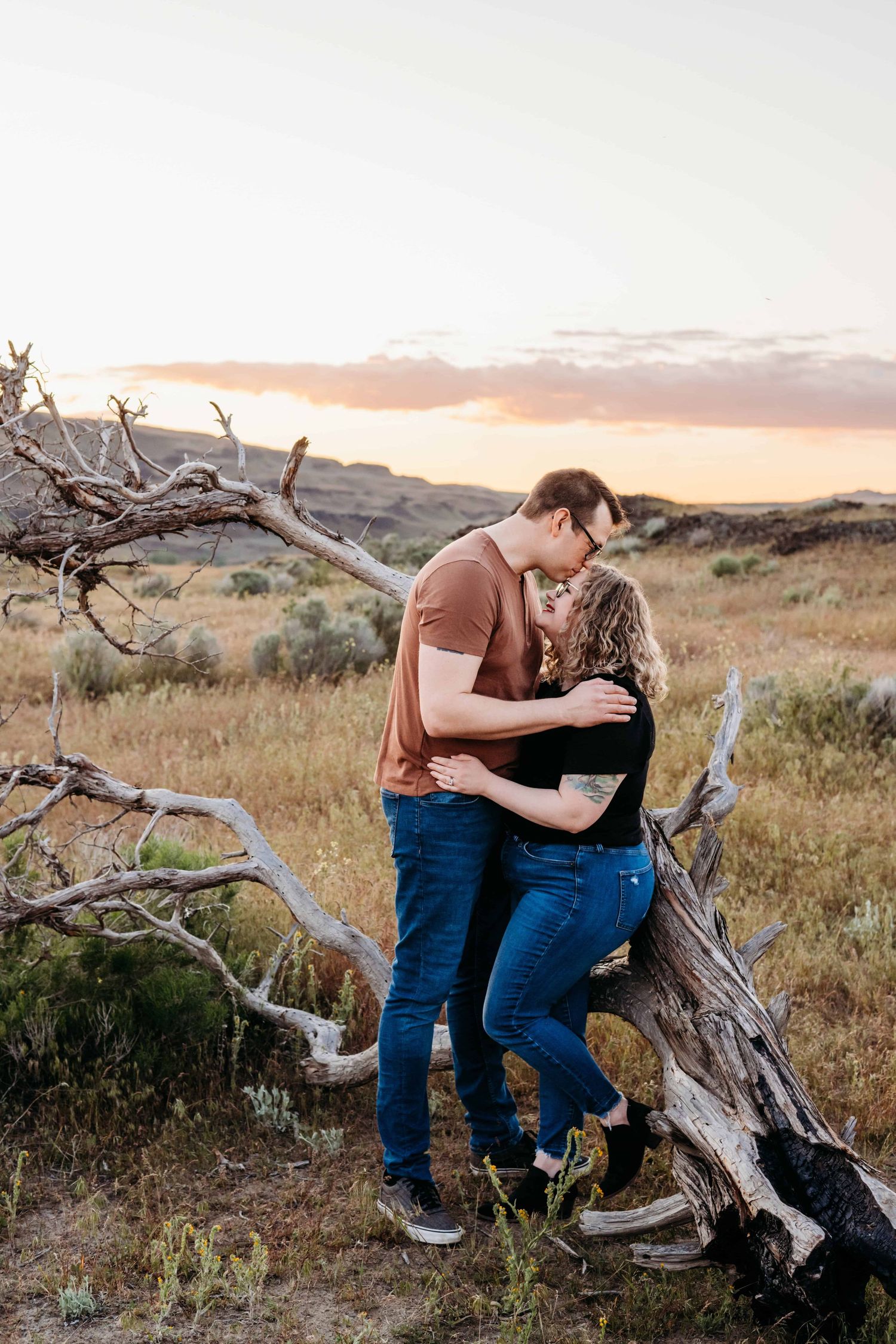 A couple leaning against a fallen tree branch while the husband kisses his wife on her forehead.