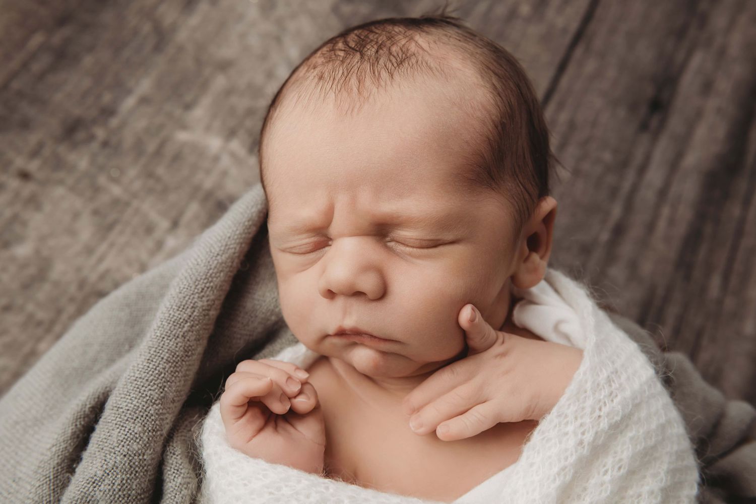 A sleeping baby laying on a gray blanket with a pensive look on his face.