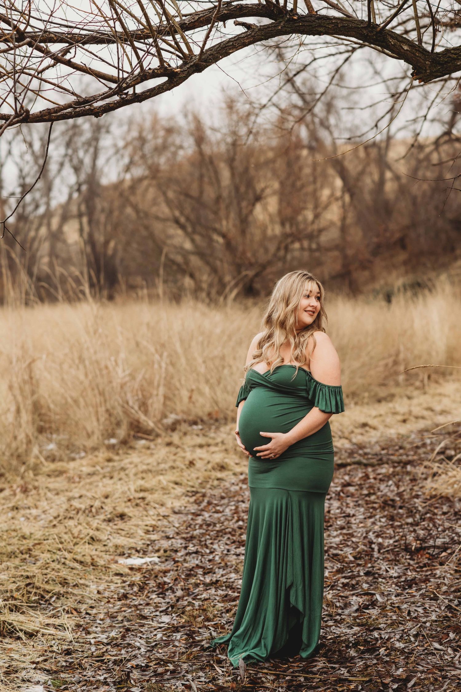 A pregnant woman standing underneath a tree branch wearing a green dress at Whitman Mission in Walla Walla, Washington.