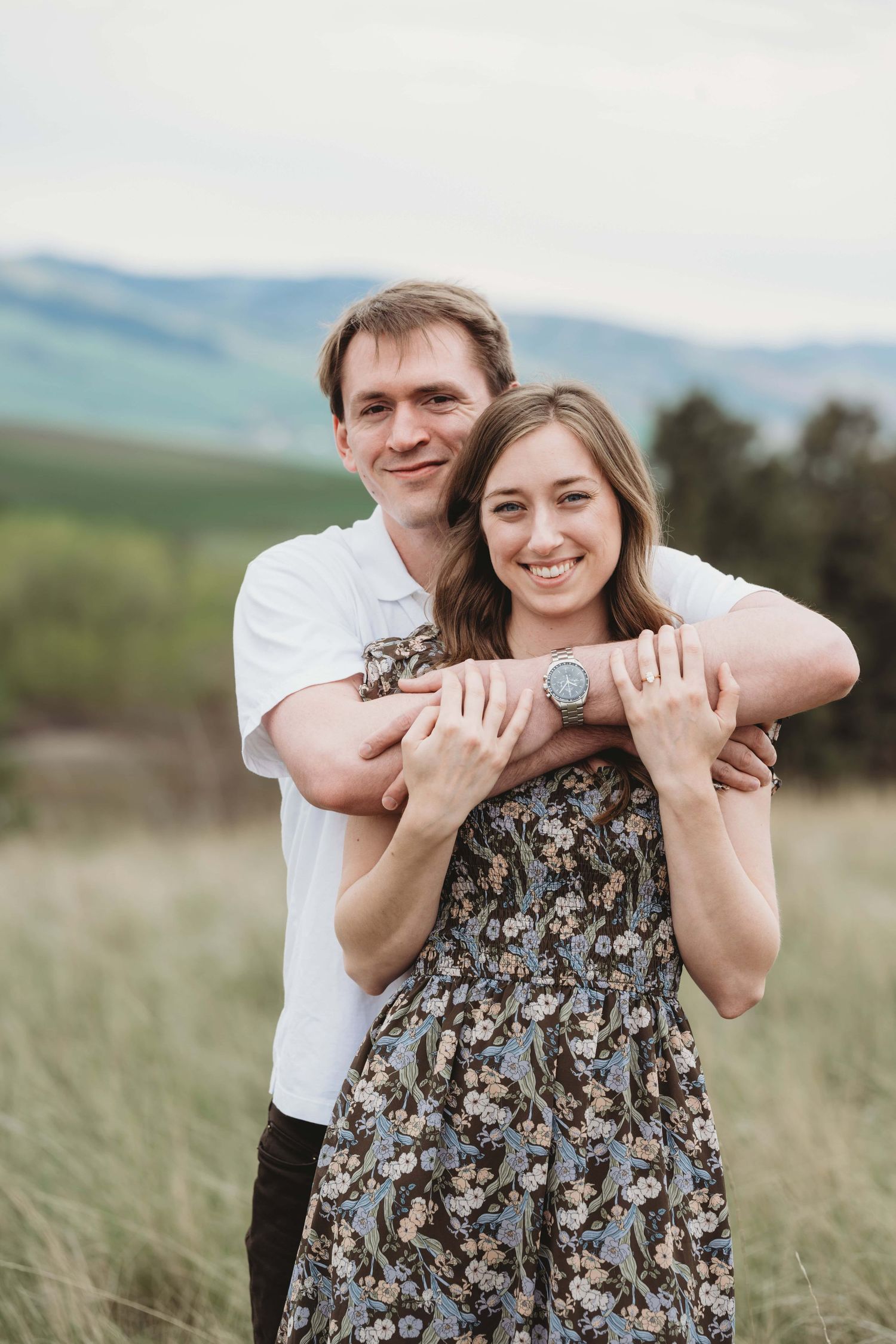 A man standing behind his new fiancee with his arms around her, smiling at the camera.