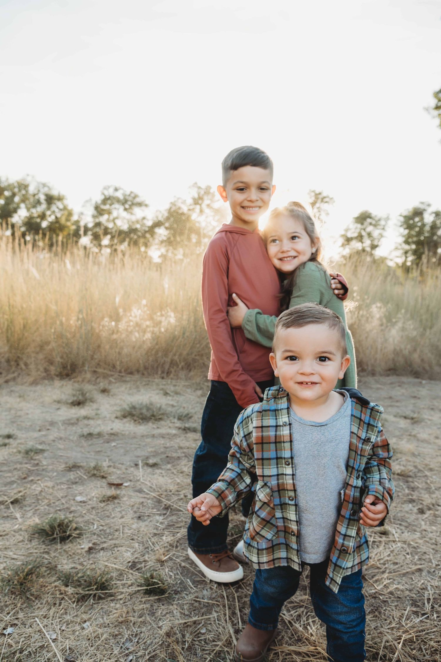 A baby boy with his two siblings in the background hugging each other.