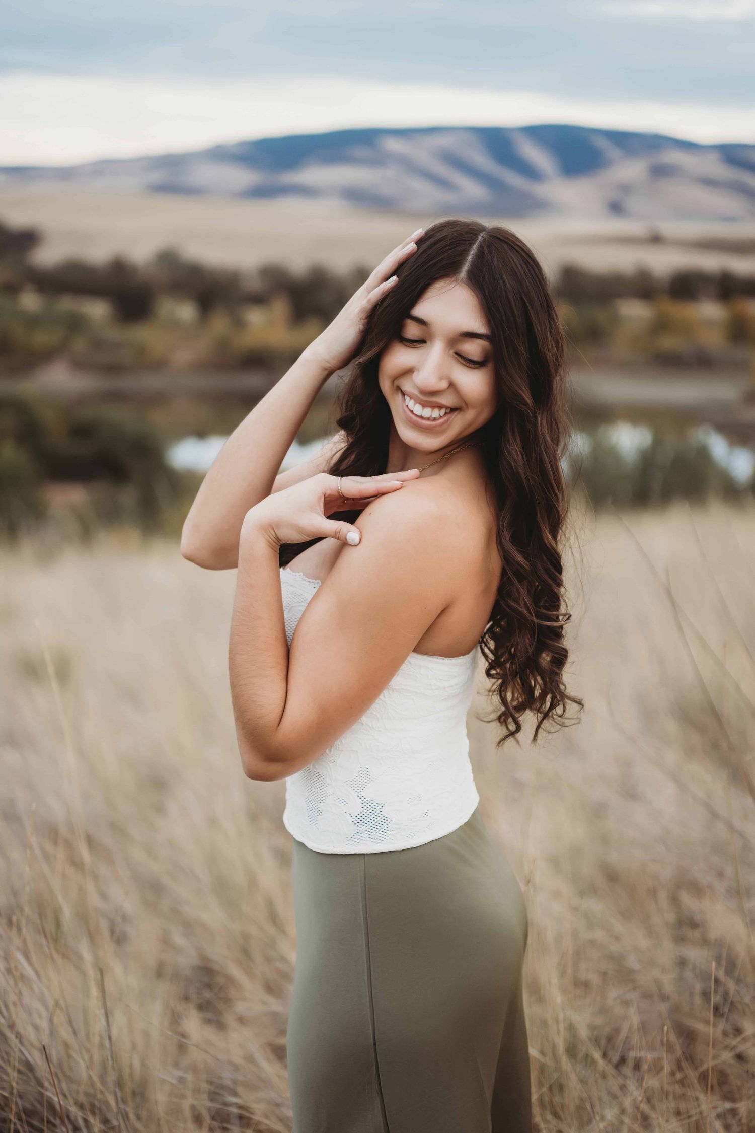 A senior wearing a white shirt and green skirt posing in front of a lake and rolling hills.