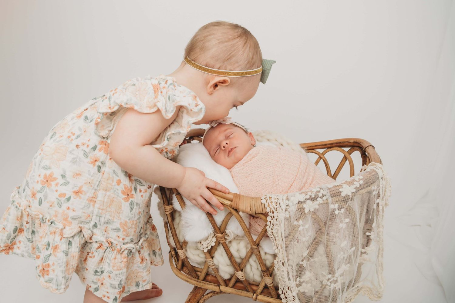 Studio portrait of a girl kissing her newborn sister as she sleeps in a bassinet.