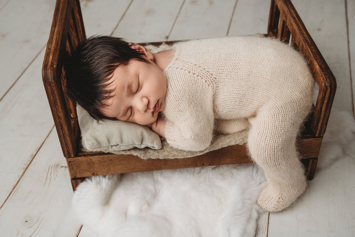 A newborn sleeping on a wooden bed with their leg hanging off of the side.