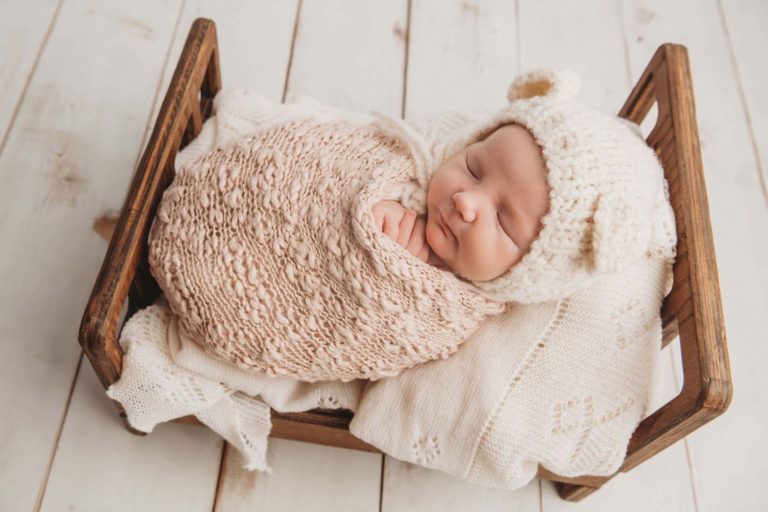 A newborn wrapped in soft pink wearing a bear bonnet and sleeping on a wooden bed.
